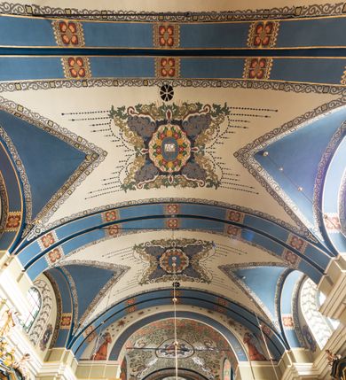 Zdjęcie nr 4: Dekoracja sklepienia nawy składa się z prostych symboli: kielicha z hostią, otwartej księgi z literami „A” i „Ω”, imienia MARIA i kotwicy. Zostały one wkomponowane w owalne medaliony w barwnej oprawie utworzonej z wieńców z żółtych i czerwonych kwiatów, złotych i zielonych wici akantów, ułożonych krzyżowo spiralnych, niebieskich ślimacznic i otaczających je stylizowanych zielonych liści akantu, między którymi przebijają się złote promienie. Owe fantazyjne kompozycje w nawie ukazane są na jasnym, kremowym tle wysklepek i niebieskich lunet i gurtów. Elementy konstrukcyjne szwów sklepiennych są podkreślone pasami z wici roślinnej. Sklepienie prezbiterium odróżnia się jaśniejszą kolorystyką. Elementy konstrukcyjne łuków podkreślają bujne czerwono-zielone kwiaty, wśród których rozpoznano piwonie. Taka sama bordiura zdobi ścianę za ołtarzem. Pośrodku umieszczono Baranka na zamkniętej księdze z chorągwią zmartwychwstania, na tle krzyża greckiego w glorii promienistej. Szwy sklepienne zaakcentowano kwiatami w układzie kandelabrowym, a wysklepki zielono-złotymi symetrycznie układającymi się wiciami roślinnymi. Na parapecie chóru muzycznego malarz przedstawił dwie sceny: „Stygmatyzacja św. Franciszka” i „Anioł grający na skrzypcach płaczącemu św. Franciszkowi”. 
