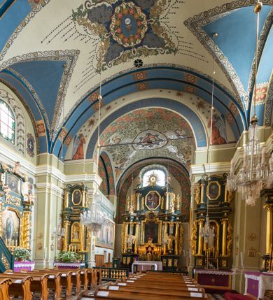 Zdjęcie nr 6: Dekoracja sklepienia nawy składa się z prostych symboli: kielicha z hostią, otwartej księgi z literami „A” i „Ω”, imienia MARIA i kotwicy. Zostały one wkomponowane w owalne medaliony w barwnej oprawie utworzonej z wieńców z żółtych i czerwonych kwiatów, złotych i zielonych wici akantów, ułożonych krzyżowo spiralnych, niebieskich ślimacznic i otaczających je stylizowanych zielonych liści akantu, między którymi przebijają się złote promienie. Owe fantazyjne kompozycje w nawie ukazane są na jasnym, kremowym tle wysklepek i niebieskich lunet i gurtów. Elementy konstrukcyjne szwów sklepiennych są podkreślone pasami z wici roślinnej. Sklepienie prezbiterium odróżnia się jaśniejszą kolorystyką. Elementy konstrukcyjne łuków podkreślają bujne czerwono-zielone kwiaty, wśród których rozpoznano piwonie. Taka sama bordiura zdobi ścianę za ołtarzem. Pośrodku umieszczono Baranka na zamkniętej księdze z chorągwią zmartwychwstania, na tle krzyża greckiego w glorii promienistej. Szwy sklepienne zaakcentowano kwiatami w układzie kandelabrowym, a wysklepki zielono-złotymi symetrycznie układającymi się wiciami roślinnymi. Na parapecie chóru muzycznego malarz przedstawił dwie sceny: „Stygmatyzacja św. Franciszka” i „Anioł grający na skrzypcach płaczącemu św. Franciszkowi”. 
