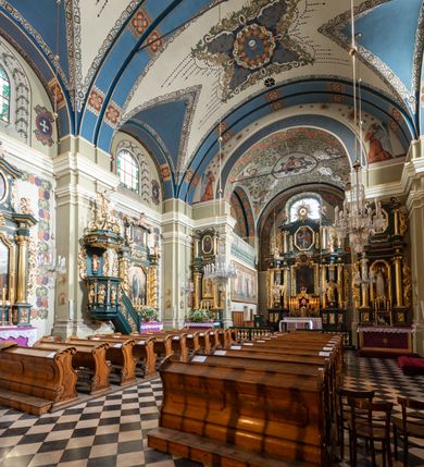 Zdjęcie nr 5: Dekoracja sklepienia nawy składa się z prostych symboli: kielicha z hostią, otwartej księgi z literami „A” i „Ω”, imienia MARIA i kotwicy. Zostały one wkomponowane w owalne medaliony w barwnej oprawie utworzonej z wieńców z żółtych i czerwonych kwiatów, złotych i zielonych wici akantów, ułożonych krzyżowo spiralnych, niebieskich ślimacznic i otaczających je stylizowanych zielonych liści akantu, między którymi przebijają się złote promienie. Owe fantazyjne kompozycje w nawie ukazane są na jasnym, kremowym tle wysklepek i niebieskich lunet i gurtów. Elementy konstrukcyjne szwów sklepiennych są podkreślone pasami z wici roślinnej. Sklepienie prezbiterium odróżnia się jaśniejszą kolorystyką. Elementy konstrukcyjne łuków podkreślają bujne czerwono-zielone kwiaty, wśród których rozpoznano piwonie. Taka sama bordiura zdobi ścianę za ołtarzem. Pośrodku umieszczono Baranka na zamkniętej księdze z chorągwią zmartwychwstania, na tle krzyża greckiego w glorii promienistej. Szwy sklepienne zaakcentowano kwiatami w układzie kandelabrowym, a wysklepki zielono-złotymi symetrycznie układającymi się wiciami roślinnymi. Na parapecie chóru muzycznego malarz przedstawił dwie sceny: „Stygmatyzacja św. Franciszka” i „Anioł grający na skrzypcach płaczącemu św. Franciszkowi”. 
