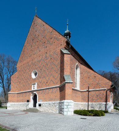 Zdjęcie nr 1: Kościół murowany z cegły, z użyciem zendrówki, na wysokim podmurowaniu z kamienia łupanego, jednonawowy, orientowany. 
Prezbiterium i nawa na planie prostokątów, nawa szersza i dłuższa od prezbiterium. Zakrystia na rzucie wydłużonego prostokąta przylega do wschodniej ściany prezbiterium, występując poza mury nawy. Prezbiterium od wschodu ujęte w narożach dwiema ukośnymi szkarpami, od południa jedną prostopadłą. Przy południowej ścianie nawy kruchta na planie kwadratu. Nawa w narożach ujęta szkarpami, od wschodu dwiema prostopadłymi, od zachodu ukośnymi. 
Prezbiterium dwuprzęsłowe, nakryte sklepieniem krzyżowo-żebrowym. Na zwornikach dwie tarcze z herbami Trzy Korony i Wieniawa. Zakrystia sklepiona kolebkowo. Nawa nakryta drewnianym stropem z kasetonami i rozetami. Kruchta sklepiona krzyżowo-żebrowo ze zwornikiem z tarczą z herbem Wieniawa. Otwór tęczowy w formie stojącego prostokąta zamkniętego łukiem ostrym. W zachodniej części nawy drewniany parapet chóru muzycznego wsparty na dwóch filarach. 
Fasada w jednej trzeciej wysokości odcięta kamiennym gzymsem kapnikowym, zakończona jodełkowym fryzem w partii muru, zwieńczona trójkątnym szczytem. Mur dekorowany zendrówką ułożoną „w zęby”, w partii szczytu w układzie rombowym. Na osi kamienny ostrołukowy portal o schodkowym nadprożu, dekorowanym krzyżującym się laskowaniem; węgary gładkie w jednej trzeciej wysokości przechodzące w piramidki i profilowanie powyżej. Nad portalem kamienna kwadratowa tablica, ujęta kamiennym obramieniem z tarczą z herbem Wieniawa. Ponad nią, na osi okulus w kamiennym obramieniu. 
Elewacje północna i południowa w części nawy w jednej trzeciej wysokości przedzielone kamiennym gzymsem kapnikowym, na zakończeniu ścian z gzymsem wieńczącym. Mur dekorowany zendrówką w układzie rombowym. Na elewacji południowej cztery okna w formie wydłużonego, stojącego prostokąta zamkniętego łukiem ostrym, w kamiennych obramieniach, podzielone laskowaniem. Południowa ściana prezbiterium zakończona gzymsem wieńczącym, w górnej części muru dekorowana zendrówką ułożoną „w zęby”. 
Szkarpy opinające nawę uskokowe, zwężające się ku górze, nakryte daszkami pulpitowymi. Szkarpy opinające prezbiterium nakryte daszkami pulpitowymi, podzielone gzymsami kapnikowymi. 
Kruchta od strony wschodniej i zachodniej podzielona kamiennym gzymsem kapnikowym, od południa zakończona gzymsem wieńczącym. Na ścianie południowej kruchty kamienny, ostrołukowy portal, o schodkowym nadprożu, dekorowanym krzyżującym się laskowaniem; węgary gładkie, w jednej trzeciej wysokości przechodzące w piramidki i profilowanie powyżej. Nad portalem kamienna tarcza z herbem Wieniawa. 
Elewacja wschodnia zwieńczona trójkątnym szczytem, w którego partii mur dekorowany jest zendrówką w układzie rombowym. Na osi okno w formie wydłużonego, stojącego prostokąta zamkniętego łukiem ostrym, w kamiennym obramieniu, zamurowane w trzech piątych wysokości, zakończone daszkiem pulpitowym. W dolnej części okna znajduje się ogrójec. 
Nawa i prezbiterium nakryte osobnymi blaszanymi dachami dwuspadowymi, kruchta i zakrystia pulpitowymi. Nad nawą blaszana wieżyczka na sygnaturkę z latarnią i hełmem baniastym.





