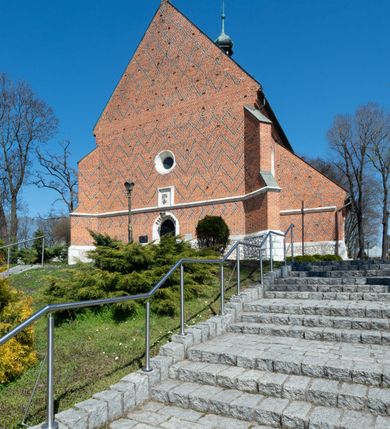 Zdjęcie nr 23: Kościół murowany z cegły, z użyciem zendrówki, na wysokim podmurowaniu z kamienia łupanego, jednonawowy, orientowany. 
Prezbiterium i nawa na planie prostokątów, nawa szersza i dłuższa od prezbiterium. Zakrystia na rzucie wydłużonego prostokąta przylega do wschodniej ściany prezbiterium, występując poza mury nawy. Prezbiterium od wschodu ujęte w narożach dwiema ukośnymi szkarpami, od południa jedną prostopadłą. Przy południowej ścianie nawy kruchta na planie kwadratu. Nawa w narożach ujęta szkarpami, od wschodu dwiema prostopadłymi, od zachodu ukośnymi. 
Prezbiterium dwuprzęsłowe, nakryte sklepieniem krzyżowo-żebrowym. Na zwornikach dwie tarcze z herbami Trzy Korony i Wieniawa. Zakrystia sklepiona kolebkowo. Nawa nakryta drewnianym stropem z kasetonami i rozetami. Kruchta sklepiona krzyżowo-żebrowo ze zwornikiem z tarczą z herbem Wieniawa. Otwór tęczowy w formie stojącego prostokąta zamkniętego łukiem ostrym. W zachodniej części nawy drewniany parapet chóru muzycznego wsparty na dwóch filarach. 
Fasada w jednej trzeciej wysokości odcięta kamiennym gzymsem kapnikowym, zakończona jodełkowym fryzem w partii muru, zwieńczona trójkątnym szczytem. Mur dekorowany zendrówką ułożoną „w zęby”, w partii szczytu w układzie rombowym. Na osi kamienny ostrołukowy portal o schodkowym nadprożu, dekorowanym krzyżującym się laskowaniem; węgary gładkie w jednej trzeciej wysokości przechodzące w piramidki i profilowanie powyżej. Nad portalem kamienna kwadratowa tablica, ujęta kamiennym obramieniem z tarczą z herbem Wieniawa. Ponad nią, na osi okulus w kamiennym obramieniu. 
Elewacje północna i południowa w części nawy w jednej trzeciej wysokości przedzielone kamiennym gzymsem kapnikowym, na zakończeniu ścian z gzymsem wieńczącym. Mur dekorowany zendrówką w układzie rombowym. Na elewacji południowej cztery okna w formie wydłużonego, stojącego prostokąta zamkniętego łukiem ostrym, w kamiennych obramieniach, podzielone laskowaniem. Południowa ściana prezbiterium zakończona gzymsem wieńczącym, w górnej części muru dekorowana zendrówką ułożoną „w zęby”. 
Szkarpy opinające nawę uskokowe, zwężające się ku górze, nakryte daszkami pulpitowymi. Szkarpy opinające prezbiterium nakryte daszkami pulpitowymi, podzielone gzymsami kapnikowymi. 
Kruchta od strony wschodniej i zachodniej podzielona kamiennym gzymsem kapnikowym, od południa zakończona gzymsem wieńczącym. Na ścianie południowej kruchty kamienny, ostrołukowy portal, o schodkowym nadprożu, dekorowanym krzyżującym się laskowaniem; węgary gładkie, w jednej trzeciej wysokości przechodzące w piramidki i profilowanie powyżej. Nad portalem kamienna tarcza z herbem Wieniawa. 
Elewacja wschodnia zwieńczona trójkątnym szczytem, w którego partii mur dekorowany jest zendrówką w układzie rombowym. Na osi okno w formie wydłużonego, stojącego prostokąta zamkniętego łukiem ostrym, w kamiennym obramieniu, zamurowane w trzech piątych wysokości, zakończone daszkiem pulpitowym. W dolnej części okna znajduje się ogrójec. 
Nawa i prezbiterium nakryte osobnymi blaszanymi dachami dwuspadowymi, kruchta i zakrystia pulpitowymi. Nad nawą blaszana wieżyczka na sygnaturkę z latarnią i hełmem baniastym.






