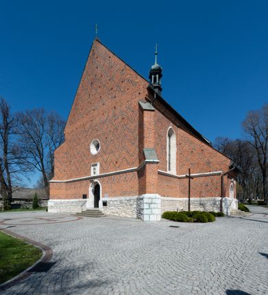 Zdjęcie nr 58: Kościół murowany z cegły, z użyciem zendrówki, na wysokim podmurowaniu z kamienia łupanego, jednonawowy, orientowany. 
Prezbiterium i nawa na planie prostokątów, nawa szersza i dłuższa od prezbiterium. Zakrystia na rzucie wydłużonego prostokąta przylega do wschodniej ściany prezbiterium, występując poza mury nawy. Prezbiterium od wschodu ujęte w narożach dwiema ukośnymi szkarpami, od południa jedną prostopadłą. Przy południowej ścianie nawy kruchta na planie kwadratu. Nawa w narożach ujęta szkarpami, od wschodu dwiema prostopadłymi, od zachodu ukośnymi. 
Prezbiterium dwuprzęsłowe, nakryte sklepieniem krzyżowo-żebrowym. Na zwornikach dwie tarcze z herbami Trzy Korony i Wieniawa. Zakrystia sklepiona kolebkowo. Nawa nakryta drewnianym stropem z kasetonami i rozetami. Kruchta sklepiona krzyżowo-żebrowo ze zwornikiem z tarczą z herbem Wieniawa. Otwór tęczowy w formie stojącego prostokąta zamkniętego łukiem ostrym. W zachodniej części nawy drewniany parapet chóru muzycznego wsparty na dwóch filarach. 
Fasada w jednej trzeciej wysokości odcięta kamiennym gzymsem kapnikowym, zakończona jodełkowym fryzem w partii muru, zwieńczona trójkątnym szczytem. Mur dekorowany zendrówką ułożoną „w zęby”, w partii szczytu w układzie rombowym. Na osi kamienny ostrołukowy portal o schodkowym nadprożu, dekorowanym krzyżującym się laskowaniem; węgary gładkie w jednej trzeciej wysokości przechodzące w piramidki i profilowanie powyżej. Nad portalem kamienna kwadratowa tablica, ujęta kamiennym obramieniem z tarczą z herbem Wieniawa. Ponad nią, na osi okulus w kamiennym obramieniu. 
Elewacje północna i południowa w części nawy w jednej trzeciej wysokości przedzielone kamiennym gzymsem kapnikowym, na zakończeniu ścian z gzymsem wieńczącym. Mur dekorowany zendrówką w układzie rombowym. Na elewacji południowej cztery okna w formie wydłużonego, stojącego prostokąta zamkniętego łukiem ostrym, w kamiennych obramieniach, podzielone laskowaniem. Południowa ściana prezbiterium zakończona gzymsem wieńczącym, w górnej części muru dekorowana zendrówką ułożoną „w zęby”. 
Szkarpy opinające nawę uskokowe, zwężające się ku górze, nakryte daszkami pulpitowymi. Szkarpy opinające prezbiterium nakryte daszkami pulpitowymi, podzielone gzymsami kapnikowymi. 
Kruchta od strony wschodniej i zachodniej podzielona kamiennym gzymsem kapnikowym, od południa zakończona gzymsem wieńczącym. Na ścianie południowej kruchty kamienny, ostrołukowy portal, o schodkowym nadprożu, dekorowanym krzyżującym się laskowaniem; węgary gładkie, w jednej trzeciej wysokości przechodzące w piramidki i profilowanie powyżej. Nad portalem kamienna tarcza z herbem Wieniawa. 
Elewacja wschodnia zwieńczona trójkątnym szczytem, w którego partii mur dekorowany jest zendrówką w układzie rombowym. Na osi okno w formie wydłużonego, stojącego prostokąta zamkniętego łukiem ostrym, w kamiennym obramieniu, zamurowane w trzech piątych wysokości, zakończone daszkiem pulpitowym. W dolnej części okna znajduje się ogrójec. 
Nawa i prezbiterium nakryte osobnymi blaszanymi dachami dwuspadowymi, kruchta i zakrystia pulpitowymi. Nad nawą blaszana wieżyczka na sygnaturkę z latarnią i hełmem baniastym.





