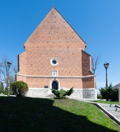Zdjęcie nr 29: Kościół murowany z cegły, z użyciem zendrówki, na wysokim podmurowaniu z kamienia łupanego, jednonawowy, orientowany. 
Prezbiterium i nawa na planie prostokątów, nawa szersza i dłuższa od prezbiterium. Zakrystia na rzucie wydłużonego prostokąta przylega do wschodniej ściany prezbiterium, występując poza mury nawy. Prezbiterium od wschodu ujęte w narożach dwiema ukośnymi szkarpami, od południa jedną prostopadłą. Przy południowej ścianie nawy kruchta na planie kwadratu. Nawa w narożach ujęta szkarpami, od wschodu dwiema prostopadłymi, od zachodu ukośnymi. 
Prezbiterium dwuprzęsłowe, nakryte sklepieniem krzyżowo-żebrowym. Na zwornikach dwie tarcze z herbami Trzy Korony i Wieniawa. Zakrystia sklepiona kolebkowo. Nawa nakryta drewnianym stropem z kasetonami i rozetami. Kruchta sklepiona krzyżowo-żebrowo ze zwornikiem z tarczą z herbem Wieniawa. Otwór tęczowy w formie stojącego prostokąta zamkniętego łukiem ostrym. W zachodniej części nawy drewniany parapet chóru muzycznego wsparty na dwóch filarach. 
Fasada w jednej trzeciej wysokości odcięta kamiennym gzymsem kapnikowym, zakończona jodełkowym fryzem w partii muru, zwieńczona trójkątnym szczytem. Mur dekorowany zendrówką ułożoną „w zęby”, w partii szczytu w układzie rombowym. Na osi kamienny ostrołukowy portal o schodkowym nadprożu, dekorowanym krzyżującym się laskowaniem; węgary gładkie w jednej trzeciej wysokości przechodzące w piramidki i profilowanie powyżej. Nad portalem kamienna kwadratowa tablica, ujęta kamiennym obramieniem z tarczą z herbem Wieniawa. Ponad nią, na osi okulus w kamiennym obramieniu. 
Elewacje północna i południowa w części nawy w jednej trzeciej wysokości przedzielone kamiennym gzymsem kapnikowym, na zakończeniu ścian z gzymsem wieńczącym. Mur dekorowany zendrówką w układzie rombowym. Na elewacji południowej cztery okna w formie wydłużonego, stojącego prostokąta zamkniętego łukiem ostrym, w kamiennych obramieniach, podzielone laskowaniem. Południowa ściana prezbiterium zakończona gzymsem wieńczącym, w górnej części muru dekorowana zendrówką ułożoną „w zęby”. 
Szkarpy opinające nawę uskokowe, zwężające się ku górze, nakryte daszkami pulpitowymi. Szkarpy opinające prezbiterium nakryte daszkami pulpitowymi, podzielone gzymsami kapnikowymi. 
Kruchta od strony wschodniej i zachodniej podzielona kamiennym gzymsem kapnikowym, od południa zakończona gzymsem wieńczącym. Na ścianie południowej kruchty kamienny, ostrołukowy portal, o schodkowym nadprożu, dekorowanym krzyżującym się laskowaniem; węgary gładkie, w jednej trzeciej wysokości przechodzące w piramidki i profilowanie powyżej. Nad portalem kamienna tarcza z herbem Wieniawa. 
Elewacja wschodnia zwieńczona trójkątnym szczytem, w którego partii mur dekorowany jest zendrówką w układzie rombowym. Na osi okno w formie wydłużonego, stojącego prostokąta zamkniętego łukiem ostrym, w kamiennym obramieniu, zamurowane w trzech piątych wysokości, zakończone daszkiem pulpitowym. W dolnej części okna znajduje się ogrójec. 
Nawa i prezbiterium nakryte osobnymi blaszanymi dachami dwuspadowymi, kruchta i zakrystia pulpitowymi. Nad nawą blaszana wieżyczka na sygnaturkę z latarnią i hełmem baniastym.





