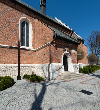 Zdjęcie nr 35: Kościół murowany z cegły, z użyciem zendrówki, na wysokim podmurowaniu z kamienia łupanego, jednonawowy, orientowany. 
Prezbiterium i nawa na planie prostokątów, nawa szersza i dłuższa od prezbiterium. Zakrystia na rzucie wydłużonego prostokąta przylega do wschodniej ściany prezbiterium, występując poza mury nawy. Prezbiterium od wschodu ujęte w narożach dwiema ukośnymi szkarpami, od południa jedną prostopadłą. Przy południowej ścianie nawy kruchta na planie kwadratu. Nawa w narożach ujęta szkarpami, od wschodu dwiema prostopadłymi, od zachodu ukośnymi. 
Prezbiterium dwuprzęsłowe, nakryte sklepieniem krzyżowo-żebrowym. Na zwornikach dwie tarcze z herbami Trzy Korony i Wieniawa. Zakrystia sklepiona kolebkowo. Nawa nakryta drewnianym stropem z kasetonami i rozetami. Kruchta sklepiona krzyżowo-żebrowo ze zwornikiem z tarczą z herbem Wieniawa. Otwór tęczowy w formie stojącego prostokąta zamkniętego łukiem ostrym. W zachodniej części nawy drewniany parapet chóru muzycznego wsparty na dwóch filarach. 
Fasada w jednej trzeciej wysokości odcięta kamiennym gzymsem kapnikowym, zakończona jodełkowym fryzem w partii muru, zwieńczona trójkątnym szczytem. Mur dekorowany zendrówką ułożoną „w zęby”, w partii szczytu w układzie rombowym. Na osi kamienny ostrołukowy portal o schodkowym nadprożu, dekorowanym krzyżującym się laskowaniem; węgary gładkie w jednej trzeciej wysokości przechodzące w piramidki i profilowanie powyżej. Nad portalem kamienna kwadratowa tablica, ujęta kamiennym obramieniem z tarczą z herbem Wieniawa. Ponad nią, na osi okulus w kamiennym obramieniu. 
Elewacje północna i południowa w części nawy w jednej trzeciej wysokości przedzielone kamiennym gzymsem kapnikowym, na zakończeniu ścian z gzymsem wieńczącym. Mur dekorowany zendrówką w układzie rombowym. Na elewacji południowej cztery okna w formie wydłużonego, stojącego prostokąta zamkniętego łukiem ostrym, w kamiennych obramieniach, podzielone laskowaniem. Południowa ściana prezbiterium zakończona gzymsem wieńczącym, w górnej części muru dekorowana zendrówką ułożoną „w zęby”. 
Szkarpy opinające nawę uskokowe, zwężające się ku górze, nakryte daszkami pulpitowymi. Szkarpy opinające prezbiterium nakryte daszkami pulpitowymi, podzielone gzymsami kapnikowymi. 
Kruchta od strony wschodniej i zachodniej podzielona kamiennym gzymsem kapnikowym, od południa zakończona gzymsem wieńczącym. Na ścianie południowej kruchty kamienny, ostrołukowy portal, o schodkowym nadprożu, dekorowanym krzyżującym się laskowaniem; węgary gładkie, w jednej trzeciej wysokości przechodzące w piramidki i profilowanie powyżej. Nad portalem kamienna tarcza z herbem Wieniawa. 
Elewacja wschodnia zwieńczona trójkątnym szczytem, w którego partii mur dekorowany jest zendrówką w układzie rombowym. Na osi okno w formie wydłużonego, stojącego prostokąta zamkniętego łukiem ostrym, w kamiennym obramieniu, zamurowane w trzech piątych wysokości, zakończone daszkiem pulpitowym. W dolnej części okna znajduje się ogrójec. 
Nawa i prezbiterium nakryte osobnymi blaszanymi dachami dwuspadowymi, kruchta i zakrystia pulpitowymi. Nad nawą blaszana wieżyczka na sygnaturkę z latarnią i hełmem baniastym.





