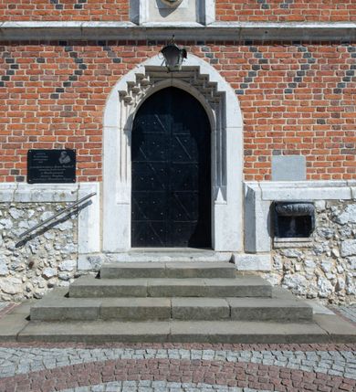 Zdjęcie nr 55: Kościół murowany z cegły, z użyciem zendrówki, na wysokim podmurowaniu z kamienia łupanego, jednonawowy, orientowany. 
Prezbiterium i nawa na planie prostokątów, nawa szersza i dłuższa od prezbiterium. Zakrystia na rzucie wydłużonego prostokąta przylega do wschodniej ściany prezbiterium, występując poza mury nawy. Prezbiterium od wschodu ujęte w narożach dwiema ukośnymi szkarpami, od południa jedną prostopadłą. Przy południowej ścianie nawy kruchta na planie kwadratu. Nawa w narożach ujęta szkarpami, od wschodu dwiema prostopadłymi, od zachodu ukośnymi. 
Prezbiterium dwuprzęsłowe, nakryte sklepieniem krzyżowo-żebrowym. Na zwornikach dwie tarcze z herbami Trzy Korony i Wieniawa. Zakrystia sklepiona kolebkowo. Nawa nakryta drewnianym stropem z kasetonami i rozetami. Kruchta sklepiona krzyżowo-żebrowo ze zwornikiem z tarczą z herbem Wieniawa. Otwór tęczowy w formie stojącego prostokąta zamkniętego łukiem ostrym. W zachodniej części nawy drewniany parapet chóru muzycznego wsparty na dwóch filarach. 
Fasada w jednej trzeciej wysokości odcięta kamiennym gzymsem kapnikowym, zakończona jodełkowym fryzem w partii muru, zwieńczona trójkątnym szczytem. Mur dekorowany zendrówką ułożoną „w zęby”, w partii szczytu w układzie rombowym. Na osi kamienny ostrołukowy portal o schodkowym nadprożu, dekorowanym krzyżującym się laskowaniem; węgary gładkie w jednej trzeciej wysokości przechodzące w piramidki i profilowanie powyżej. Nad portalem kamienna kwadratowa tablica, ujęta kamiennym obramieniem z tarczą z herbem Wieniawa. Ponad nią, na osi okulus w kamiennym obramieniu. 
Elewacje północna i południowa w części nawy w jednej trzeciej wysokości przedzielone kamiennym gzymsem kapnikowym, na zakończeniu ścian z gzymsem wieńczącym. Mur dekorowany zendrówką w układzie rombowym. Na elewacji południowej cztery okna w formie wydłużonego, stojącego prostokąta zamkniętego łukiem ostrym, w kamiennych obramieniach, podzielone laskowaniem. Południowa ściana prezbiterium zakończona gzymsem wieńczącym, w górnej części muru dekorowana zendrówką ułożoną „w zęby”. 
Szkarpy opinające nawę uskokowe, zwężające się ku górze, nakryte daszkami pulpitowymi. Szkarpy opinające prezbiterium nakryte daszkami pulpitowymi, podzielone gzymsami kapnikowymi. 
Kruchta od strony wschodniej i zachodniej podzielona kamiennym gzymsem kapnikowym, od południa zakończona gzymsem wieńczącym. Na ścianie południowej kruchty kamienny, ostrołukowy portal, o schodkowym nadprożu, dekorowanym krzyżującym się laskowaniem; węgary gładkie, w jednej trzeciej wysokości przechodzące w piramidki i profilowanie powyżej. Nad portalem kamienna tarcza z herbem Wieniawa. 
Elewacja wschodnia zwieńczona trójkątnym szczytem, w którego partii mur dekorowany jest zendrówką w układzie rombowym. Na osi okno w formie wydłużonego, stojącego prostokąta zamkniętego łukiem ostrym, w kamiennym obramieniu, zamurowane w trzech piątych wysokości, zakończone daszkiem pulpitowym. W dolnej części okna znajduje się ogrójec. 
Nawa i prezbiterium nakryte osobnymi blaszanymi dachami dwuspadowymi, kruchta i zakrystia pulpitowymi. Nad nawą blaszana wieżyczka na sygnaturkę z latarnią i hełmem baniastym.





