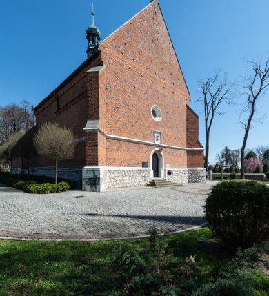 Zdjęcie nr 68: Kościół murowany z cegły, z użyciem zendrówki, na wysokim podmurowaniu z kamienia łupanego, jednonawowy, orientowany. 
Prezbiterium i nawa na planie prostokątów, nawa szersza i dłuższa od prezbiterium. Zakrystia na rzucie wydłużonego prostokąta przylega do wschodniej ściany prezbiterium, występując poza mury nawy. Prezbiterium od wschodu ujęte w narożach dwiema ukośnymi szkarpami, od południa jedną prostopadłą. Przy południowej ścianie nawy kruchta na planie kwadratu. Nawa w narożach ujęta szkarpami, od wschodu dwiema prostopadłymi, od zachodu ukośnymi. 
Prezbiterium dwuprzęsłowe, nakryte sklepieniem krzyżowo-żebrowym. Na zwornikach dwie tarcze z herbami Trzy Korony i Wieniawa. Zakrystia sklepiona kolebkowo. Nawa nakryta drewnianym stropem z kasetonami i rozetami. Kruchta sklepiona krzyżowo-żebrowo ze zwornikiem z tarczą z herbem Wieniawa. Otwór tęczowy w formie stojącego prostokąta zamkniętego łukiem ostrym. W zachodniej części nawy drewniany parapet chóru muzycznego wsparty na dwóch filarach. 
Fasada w jednej trzeciej wysokości odcięta kamiennym gzymsem kapnikowym, zakończona jodełkowym fryzem w partii muru, zwieńczona trójkątnym szczytem. Mur dekorowany zendrówką ułożoną „w zęby”, w partii szczytu w układzie rombowym. Na osi kamienny ostrołukowy portal o schodkowym nadprożu, dekorowanym krzyżującym się laskowaniem; węgary gładkie w jednej trzeciej wysokości przechodzące w piramidki i profilowanie powyżej. Nad portalem kamienna kwadratowa tablica, ujęta kamiennym obramieniem z tarczą z herbem Wieniawa. Ponad nią, na osi okulus w kamiennym obramieniu. 
Elewacje północna i południowa w części nawy w jednej trzeciej wysokości przedzielone kamiennym gzymsem kapnikowym, na zakończeniu ścian z gzymsem wieńczącym. Mur dekorowany zendrówką w układzie rombowym. Na elewacji południowej cztery okna w formie wydłużonego, stojącego prostokąta zamkniętego łukiem ostrym, w kamiennych obramieniach, podzielone laskowaniem. Południowa ściana prezbiterium zakończona gzymsem wieńczącym, w górnej części muru dekorowana zendrówką ułożoną „w zęby”. 
Szkarpy opinające nawę uskokowe, zwężające się ku górze, nakryte daszkami pulpitowymi. Szkarpy opinające prezbiterium nakryte daszkami pulpitowymi, podzielone gzymsami kapnikowymi. 
Kruchta od strony wschodniej i zachodniej podzielona kamiennym gzymsem kapnikowym, od południa zakończona gzymsem wieńczącym. Na ścianie południowej kruchty kamienny, ostrołukowy portal, o schodkowym nadprożu, dekorowanym krzyżującym się laskowaniem; węgary gładkie, w jednej trzeciej wysokości przechodzące w piramidki i profilowanie powyżej. Nad portalem kamienna tarcza z herbem Wieniawa. 
Elewacja wschodnia zwieńczona trójkątnym szczytem, w którego partii mur dekorowany jest zendrówką w układzie rombowym. Na osi okno w formie wydłużonego, stojącego prostokąta zamkniętego łukiem ostrym, w kamiennym obramieniu, zamurowane w trzech piątych wysokości, zakończone daszkiem pulpitowym. W dolnej części okna znajduje się ogrójec. 
Nawa i prezbiterium nakryte osobnymi blaszanymi dachami dwuspadowymi, kruchta i zakrystia pulpitowymi. Nad nawą blaszana wieżyczka na sygnaturkę z latarnią i hełmem baniastym.





