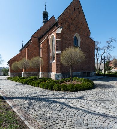 Zdjęcie nr 39: Kościół murowany z cegły, z użyciem zendrówki, na wysokim podmurowaniu z kamienia łupanego, jednonawowy, orientowany. 
Prezbiterium i nawa na planie prostokątów, nawa szersza i dłuższa od prezbiterium. Zakrystia na rzucie wydłużonego prostokąta przylega do wschodniej ściany prezbiterium, występując poza mury nawy. Prezbiterium od wschodu ujęte w narożach dwiema ukośnymi szkarpami, od południa jedną prostopadłą. Przy południowej ścianie nawy kruchta na planie kwadratu. Nawa w narożach ujęta szkarpami, od wschodu dwiema prostopadłymi, od zachodu ukośnymi. 
Prezbiterium dwuprzęsłowe, nakryte sklepieniem krzyżowo-żebrowym. Na zwornikach dwie tarcze z herbami Trzy Korony i Wieniawa. Zakrystia sklepiona kolebkowo. Nawa nakryta drewnianym stropem z kasetonami i rozetami. Kruchta sklepiona krzyżowo-żebrowo ze zwornikiem z tarczą z herbem Wieniawa. Otwór tęczowy w formie stojącego prostokąta zamkniętego łukiem ostrym. W zachodniej części nawy drewniany parapet chóru muzycznego wsparty na dwóch filarach. 
Fasada w jednej trzeciej wysokości odcięta kamiennym gzymsem kapnikowym, zakończona jodełkowym fryzem w partii muru, zwieńczona trójkątnym szczytem. Mur dekorowany zendrówką ułożoną „w zęby”, w partii szczytu w układzie rombowym. Na osi kamienny ostrołukowy portal o schodkowym nadprożu, dekorowanym krzyżującym się laskowaniem; węgary gładkie w jednej trzeciej wysokości przechodzące w piramidki i profilowanie powyżej. Nad portalem kamienna kwadratowa tablica, ujęta kamiennym obramieniem z tarczą z herbem Wieniawa. Ponad nią, na osi okulus w kamiennym obramieniu. 
Elewacje północna i południowa w części nawy w jednej trzeciej wysokości przedzielone kamiennym gzymsem kapnikowym, na zakończeniu ścian z gzymsem wieńczącym. Mur dekorowany zendrówką w układzie rombowym. Na elewacji południowej cztery okna w formie wydłużonego, stojącego prostokąta zamkniętego łukiem ostrym, w kamiennych obramieniach, podzielone laskowaniem. Południowa ściana prezbiterium zakończona gzymsem wieńczącym, w górnej części muru dekorowana zendrówką ułożoną „w zęby”. 
Szkarpy opinające nawę uskokowe, zwężające się ku górze, nakryte daszkami pulpitowymi. Szkarpy opinające prezbiterium nakryte daszkami pulpitowymi, podzielone gzymsami kapnikowymi. 
Kruchta od strony wschodniej i zachodniej podzielona kamiennym gzymsem kapnikowym, od południa zakończona gzymsem wieńczącym. Na ścianie południowej kruchty kamienny, ostrołukowy portal, o schodkowym nadprożu, dekorowanym krzyżującym się laskowaniem; węgary gładkie, w jednej trzeciej wysokości przechodzące w piramidki i profilowanie powyżej. Nad portalem kamienna tarcza z herbem Wieniawa. 
Elewacja wschodnia zwieńczona trójkątnym szczytem, w którego partii mur dekorowany jest zendrówką w układzie rombowym. Na osi okno w formie wydłużonego, stojącego prostokąta zamkniętego łukiem ostrym, w kamiennym obramieniu, zamurowane w trzech piątych wysokości, zakończone daszkiem pulpitowym. W dolnej części okna znajduje się ogrójec. 
Nawa i prezbiterium nakryte osobnymi blaszanymi dachami dwuspadowymi, kruchta i zakrystia pulpitowymi. Nad nawą blaszana wieżyczka na sygnaturkę z latarnią i hełmem baniastym.





