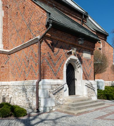 Zdjęcie nr 53: Kościół murowany z cegły, z użyciem zendrówki, na wysokim podmurowaniu z kamienia łupanego, jednonawowy, orientowany. 
Prezbiterium i nawa na planie prostokątów, nawa szersza i dłuższa od prezbiterium. Zakrystia na rzucie wydłużonego prostokąta przylega do wschodniej ściany prezbiterium, występując poza mury nawy. Prezbiterium od wschodu ujęte w narożach dwiema ukośnymi szkarpami, od południa jedną prostopadłą. Przy południowej ścianie nawy kruchta na planie kwadratu. Nawa w narożach ujęta szkarpami, od wschodu dwiema prostopadłymi, od zachodu ukośnymi. 
Prezbiterium dwuprzęsłowe, nakryte sklepieniem krzyżowo-żebrowym. Na zwornikach dwie tarcze z herbami Trzy Korony i Wieniawa. Zakrystia sklepiona kolebkowo. Nawa nakryta drewnianym stropem z kasetonami i rozetami. Kruchta sklepiona krzyżowo-żebrowo ze zwornikiem z tarczą z herbem Wieniawa. Otwór tęczowy w formie stojącego prostokąta zamkniętego łukiem ostrym. W zachodniej części nawy drewniany parapet chóru muzycznego wsparty na dwóch filarach. 
Fasada w jednej trzeciej wysokości odcięta kamiennym gzymsem kapnikowym, zakończona jodełkowym fryzem w partii muru, zwieńczona trójkątnym szczytem. Mur dekorowany zendrówką ułożoną „w zęby”, w partii szczytu w układzie rombowym. Na osi kamienny ostrołukowy portal o schodkowym nadprożu, dekorowanym krzyżującym się laskowaniem; węgary gładkie w jednej trzeciej wysokości przechodzące w piramidki i profilowanie powyżej. Nad portalem kamienna kwadratowa tablica, ujęta kamiennym obramieniem z tarczą z herbem Wieniawa. Ponad nią, na osi okulus w kamiennym obramieniu. 
Elewacje północna i południowa w części nawy w jednej trzeciej wysokości przedzielone kamiennym gzymsem kapnikowym, na zakończeniu ścian z gzymsem wieńczącym. Mur dekorowany zendrówką w układzie rombowym. Na elewacji południowej cztery okna w formie wydłużonego, stojącego prostokąta zamkniętego łukiem ostrym, w kamiennych obramieniach, podzielone laskowaniem. Południowa ściana prezbiterium zakończona gzymsem wieńczącym, w górnej części muru dekorowana zendrówką ułożoną „w zęby”. 
Szkarpy opinające nawę uskokowe, zwężające się ku górze, nakryte daszkami pulpitowymi. Szkarpy opinające prezbiterium nakryte daszkami pulpitowymi, podzielone gzymsami kapnikowymi. 
Kruchta od strony wschodniej i zachodniej podzielona kamiennym gzymsem kapnikowym, od południa zakończona gzymsem wieńczącym. Na ścianie południowej kruchty kamienny, ostrołukowy portal, o schodkowym nadprożu, dekorowanym krzyżującym się laskowaniem; węgary gładkie, w jednej trzeciej wysokości przechodzące w piramidki i profilowanie powyżej. Nad portalem kamienna tarcza z herbem Wieniawa. 
Elewacja wschodnia zwieńczona trójkątnym szczytem, w którego partii mur dekorowany jest zendrówką w układzie rombowym. Na osi okno w formie wydłużonego, stojącego prostokąta zamkniętego łukiem ostrym, w kamiennym obramieniu, zamurowane w trzech piątych wysokości, zakończone daszkiem pulpitowym. W dolnej części okna znajduje się ogrójec. 
Nawa i prezbiterium nakryte osobnymi blaszanymi dachami dwuspadowymi, kruchta i zakrystia pulpitowymi. Nad nawą blaszana wieżyczka na sygnaturkę z latarnią i hełmem baniastym.





