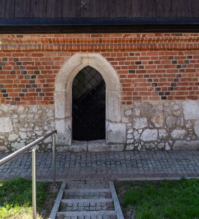 Zdjęcie nr 65: Kościół murowany z cegły, z użyciem zendrówki, na wysokim podmurowaniu z kamienia łupanego, jednonawowy, orientowany. 
Prezbiterium i nawa na planie prostokątów, nawa szersza i dłuższa od prezbiterium. Zakrystia na rzucie wydłużonego prostokąta przylega do wschodniej ściany prezbiterium, występując poza mury nawy. Prezbiterium od wschodu ujęte w narożach dwiema ukośnymi szkarpami, od południa jedną prostopadłą. Przy południowej ścianie nawy kruchta na planie kwadratu. Nawa w narożach ujęta szkarpami, od wschodu dwiema prostopadłymi, od zachodu ukośnymi. 
Prezbiterium dwuprzęsłowe, nakryte sklepieniem krzyżowo-żebrowym. Na zwornikach dwie tarcze z herbami Trzy Korony i Wieniawa. Zakrystia sklepiona kolebkowo. Nawa nakryta drewnianym stropem z kasetonami i rozetami. Kruchta sklepiona krzyżowo-żebrowo ze zwornikiem z tarczą z herbem Wieniawa. Otwór tęczowy w formie stojącego prostokąta zamkniętego łukiem ostrym. W zachodniej części nawy drewniany parapet chóru muzycznego wsparty na dwóch filarach. 
Fasada w jednej trzeciej wysokości odcięta kamiennym gzymsem kapnikowym, zakończona jodełkowym fryzem w partii muru, zwieńczona trójkątnym szczytem. Mur dekorowany zendrówką ułożoną „w zęby”, w partii szczytu w układzie rombowym. Na osi kamienny ostrołukowy portal o schodkowym nadprożu, dekorowanym krzyżującym się laskowaniem; węgary gładkie w jednej trzeciej wysokości przechodzące w piramidki i profilowanie powyżej. Nad portalem kamienna kwadratowa tablica, ujęta kamiennym obramieniem z tarczą z herbem Wieniawa. Ponad nią, na osi okulus w kamiennym obramieniu. 
Elewacje północna i południowa w części nawy w jednej trzeciej wysokości przedzielone kamiennym gzymsem kapnikowym, na zakończeniu ścian z gzymsem wieńczącym. Mur dekorowany zendrówką w układzie rombowym. Na elewacji południowej cztery okna w formie wydłużonego, stojącego prostokąta zamkniętego łukiem ostrym, w kamiennych obramieniach, podzielone laskowaniem. Południowa ściana prezbiterium zakończona gzymsem wieńczącym, w górnej części muru dekorowana zendrówką ułożoną „w zęby”. 
Szkarpy opinające nawę uskokowe, zwężające się ku górze, nakryte daszkami pulpitowymi. Szkarpy opinające prezbiterium nakryte daszkami pulpitowymi, podzielone gzymsami kapnikowymi. 
Kruchta od strony wschodniej i zachodniej podzielona kamiennym gzymsem kapnikowym, od południa zakończona gzymsem wieńczącym. Na ścianie południowej kruchty kamienny, ostrołukowy portal, o schodkowym nadprożu, dekorowanym krzyżującym się laskowaniem; węgary gładkie, w jednej trzeciej wysokości przechodzące w piramidki i profilowanie powyżej. Nad portalem kamienna tarcza z herbem Wieniawa. 
Elewacja wschodnia zwieńczona trójkątnym szczytem, w którego partii mur dekorowany jest zendrówką w układzie rombowym. Na osi okno w formie wydłużonego, stojącego prostokąta zamkniętego łukiem ostrym, w kamiennym obramieniu, zamurowane w trzech piątych wysokości, zakończone daszkiem pulpitowym. W dolnej części okna znajduje się ogrójec. 
Nawa i prezbiterium nakryte osobnymi blaszanymi dachami dwuspadowymi, kruchta i zakrystia pulpitowymi. Nad nawą blaszana wieżyczka na sygnaturkę z latarnią i hełmem baniastym.





