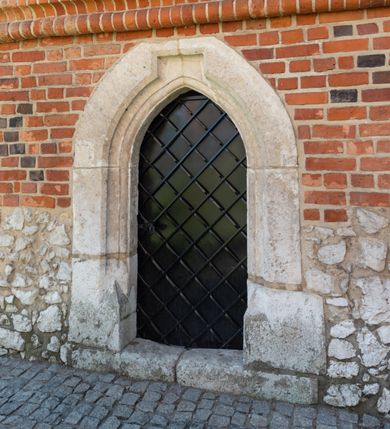 Zdjęcie nr 63: Kościół murowany z cegły, z użyciem zendrówki, na wysokim podmurowaniu z kamienia łupanego, jednonawowy, orientowany. 
Prezbiterium i nawa na planie prostokątów, nawa szersza i dłuższa od prezbiterium. Zakrystia na rzucie wydłużonego prostokąta przylega do wschodniej ściany prezbiterium, występując poza mury nawy. Prezbiterium od wschodu ujęte w narożach dwiema ukośnymi szkarpami, od południa jedną prostopadłą. Przy południowej ścianie nawy kruchta na planie kwadratu. Nawa w narożach ujęta szkarpami, od wschodu dwiema prostopadłymi, od zachodu ukośnymi. 
Prezbiterium dwuprzęsłowe, nakryte sklepieniem krzyżowo-żebrowym. Na zwornikach dwie tarcze z herbami Trzy Korony i Wieniawa. Zakrystia sklepiona kolebkowo. Nawa nakryta drewnianym stropem z kasetonami i rozetami. Kruchta sklepiona krzyżowo-żebrowo ze zwornikiem z tarczą z herbem Wieniawa. Otwór tęczowy w formie stojącego prostokąta zamkniętego łukiem ostrym. W zachodniej części nawy drewniany parapet chóru muzycznego wsparty na dwóch filarach. 
Fasada w jednej trzeciej wysokości odcięta kamiennym gzymsem kapnikowym, zakończona jodełkowym fryzem w partii muru, zwieńczona trójkątnym szczytem. Mur dekorowany zendrówką ułożoną „w zęby”, w partii szczytu w układzie rombowym. Na osi kamienny ostrołukowy portal o schodkowym nadprożu, dekorowanym krzyżującym się laskowaniem; węgary gładkie w jednej trzeciej wysokości przechodzące w piramidki i profilowanie powyżej. Nad portalem kamienna kwadratowa tablica, ujęta kamiennym obramieniem z tarczą z herbem Wieniawa. Ponad nią, na osi okulus w kamiennym obramieniu. 
Elewacje północna i południowa w części nawy w jednej trzeciej wysokości przedzielone kamiennym gzymsem kapnikowym, na zakończeniu ścian z gzymsem wieńczącym. Mur dekorowany zendrówką w układzie rombowym. Na elewacji południowej cztery okna w formie wydłużonego, stojącego prostokąta zamkniętego łukiem ostrym, w kamiennych obramieniach, podzielone laskowaniem. Południowa ściana prezbiterium zakończona gzymsem wieńczącym, w górnej części muru dekorowana zendrówką ułożoną „w zęby”. 
Szkarpy opinające nawę uskokowe, zwężające się ku górze, nakryte daszkami pulpitowymi. Szkarpy opinające prezbiterium nakryte daszkami pulpitowymi, podzielone gzymsami kapnikowymi. 
Kruchta od strony wschodniej i zachodniej podzielona kamiennym gzymsem kapnikowym, od południa zakończona gzymsem wieńczącym. Na ścianie południowej kruchty kamienny, ostrołukowy portal, o schodkowym nadprożu, dekorowanym krzyżującym się laskowaniem; węgary gładkie, w jednej trzeciej wysokości przechodzące w piramidki i profilowanie powyżej. Nad portalem kamienna tarcza z herbem Wieniawa. 
Elewacja wschodnia zwieńczona trójkątnym szczytem, w którego partii mur dekorowany jest zendrówką w układzie rombowym. Na osi okno w formie wydłużonego, stojącego prostokąta zamkniętego łukiem ostrym, w kamiennym obramieniu, zamurowane w trzech piątych wysokości, zakończone daszkiem pulpitowym. W dolnej części okna znajduje się ogrójec. 
Nawa i prezbiterium nakryte osobnymi blaszanymi dachami dwuspadowymi, kruchta i zakrystia pulpitowymi. Nad nawą blaszana wieżyczka na sygnaturkę z latarnią i hełmem baniastym.





