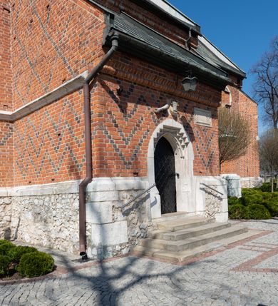 Zdjęcie nr 59: Kościół murowany z cegły, z użyciem zendrówki, na wysokim podmurowaniu z kamienia łupanego, jednonawowy, orientowany. 
Prezbiterium i nawa na planie prostokątów, nawa szersza i dłuższa od prezbiterium. Zakrystia na rzucie wydłużonego prostokąta przylega do wschodniej ściany prezbiterium, występując poza mury nawy. Prezbiterium od wschodu ujęte w narożach dwiema ukośnymi szkarpami, od południa jedną prostopadłą. Przy południowej ścianie nawy kruchta na planie kwadratu. Nawa w narożach ujęta szkarpami, od wschodu dwiema prostopadłymi, od zachodu ukośnymi. 
Prezbiterium dwuprzęsłowe, nakryte sklepieniem krzyżowo-żebrowym. Na zwornikach dwie tarcze z herbami Trzy Korony i Wieniawa. Zakrystia sklepiona kolebkowo. Nawa nakryta drewnianym stropem z kasetonami i rozetami. Kruchta sklepiona krzyżowo-żebrowo ze zwornikiem z tarczą z herbem Wieniawa. Otwór tęczowy w formie stojącego prostokąta zamkniętego łukiem ostrym. W zachodniej części nawy drewniany parapet chóru muzycznego wsparty na dwóch filarach. 
Fasada w jednej trzeciej wysokości odcięta kamiennym gzymsem kapnikowym, zakończona jodełkowym fryzem w partii muru, zwieńczona trójkątnym szczytem. Mur dekorowany zendrówką ułożoną „w zęby”, w partii szczytu w układzie rombowym. Na osi kamienny ostrołukowy portal o schodkowym nadprożu, dekorowanym krzyżującym się laskowaniem; węgary gładkie w jednej trzeciej wysokości przechodzące w piramidki i profilowanie powyżej. Nad portalem kamienna kwadratowa tablica, ujęta kamiennym obramieniem z tarczą z herbem Wieniawa. Ponad nią, na osi okulus w kamiennym obramieniu. 
Elewacje północna i południowa w części nawy w jednej trzeciej wysokości przedzielone kamiennym gzymsem kapnikowym, na zakończeniu ścian z gzymsem wieńczącym. Mur dekorowany zendrówką w układzie rombowym. Na elewacji południowej cztery okna w formie wydłużonego, stojącego prostokąta zamkniętego łukiem ostrym, w kamiennych obramieniach, podzielone laskowaniem. Południowa ściana prezbiterium zakończona gzymsem wieńczącym, w górnej części muru dekorowana zendrówką ułożoną „w zęby”. 
Szkarpy opinające nawę uskokowe, zwężające się ku górze, nakryte daszkami pulpitowymi. Szkarpy opinające prezbiterium nakryte daszkami pulpitowymi, podzielone gzymsami kapnikowymi. 
Kruchta od strony wschodniej i zachodniej podzielona kamiennym gzymsem kapnikowym, od południa zakończona gzymsem wieńczącym. Na ścianie południowej kruchty kamienny, ostrołukowy portal, o schodkowym nadprożu, dekorowanym krzyżującym się laskowaniem; węgary gładkie, w jednej trzeciej wysokości przechodzące w piramidki i profilowanie powyżej. Nad portalem kamienna tarcza z herbem Wieniawa. 
Elewacja wschodnia zwieńczona trójkątnym szczytem, w którego partii mur dekorowany jest zendrówką w układzie rombowym. Na osi okno w formie wydłużonego, stojącego prostokąta zamkniętego łukiem ostrym, w kamiennym obramieniu, zamurowane w trzech piątych wysokości, zakończone daszkiem pulpitowym. W dolnej części okna znajduje się ogrójec. 
Nawa i prezbiterium nakryte osobnymi blaszanymi dachami dwuspadowymi, kruchta i zakrystia pulpitowymi. Nad nawą blaszana wieżyczka na sygnaturkę z latarnią i hełmem baniastym.





