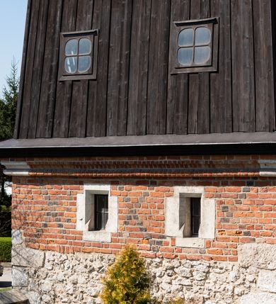 Zdjęcie nr 52: Kościół murowany z cegły, z użyciem zendrówki, na wysokim podmurowaniu z kamienia łupanego, jednonawowy, orientowany. 
Prezbiterium i nawa na planie prostokątów, nawa szersza i dłuższa od prezbiterium. Zakrystia na rzucie wydłużonego prostokąta przylega do wschodniej ściany prezbiterium, występując poza mury nawy. Prezbiterium od wschodu ujęte w narożach dwiema ukośnymi szkarpami, od południa jedną prostopadłą. Przy południowej ścianie nawy kruchta na planie kwadratu. Nawa w narożach ujęta szkarpami, od wschodu dwiema prostopadłymi, od zachodu ukośnymi. 
Prezbiterium dwuprzęsłowe, nakryte sklepieniem krzyżowo-żebrowym. Na zwornikach dwie tarcze z herbami Trzy Korony i Wieniawa. Zakrystia sklepiona kolebkowo. Nawa nakryta drewnianym stropem z kasetonami i rozetami. Kruchta sklepiona krzyżowo-żebrowo ze zwornikiem z tarczą z herbem Wieniawa. Otwór tęczowy w formie stojącego prostokąta zamkniętego łukiem ostrym. W zachodniej części nawy drewniany parapet chóru muzycznego wsparty na dwóch filarach. 
Fasada w jednej trzeciej wysokości odcięta kamiennym gzymsem kapnikowym, zakończona jodełkowym fryzem w partii muru, zwieńczona trójkątnym szczytem. Mur dekorowany zendrówką ułożoną „w zęby”, w partii szczytu w układzie rombowym. Na osi kamienny ostrołukowy portal o schodkowym nadprożu, dekorowanym krzyżującym się laskowaniem; węgary gładkie w jednej trzeciej wysokości przechodzące w piramidki i profilowanie powyżej. Nad portalem kamienna kwadratowa tablica, ujęta kamiennym obramieniem z tarczą z herbem Wieniawa. Ponad nią, na osi okulus w kamiennym obramieniu. 
Elewacje północna i południowa w części nawy w jednej trzeciej wysokości przedzielone kamiennym gzymsem kapnikowym, na zakończeniu ścian z gzymsem wieńczącym. Mur dekorowany zendrówką w układzie rombowym. Na elewacji południowej cztery okna w formie wydłużonego, stojącego prostokąta zamkniętego łukiem ostrym, w kamiennych obramieniach, podzielone laskowaniem. Południowa ściana prezbiterium zakończona gzymsem wieńczącym, w górnej części muru dekorowana zendrówką ułożoną „w zęby”. 
Szkarpy opinające nawę uskokowe, zwężające się ku górze, nakryte daszkami pulpitowymi. Szkarpy opinające prezbiterium nakryte daszkami pulpitowymi, podzielone gzymsami kapnikowymi. 
Kruchta od strony wschodniej i zachodniej podzielona kamiennym gzymsem kapnikowym, od południa zakończona gzymsem wieńczącym. Na ścianie południowej kruchty kamienny, ostrołukowy portal, o schodkowym nadprożu, dekorowanym krzyżującym się laskowaniem; węgary gładkie, w jednej trzeciej wysokości przechodzące w piramidki i profilowanie powyżej. Nad portalem kamienna tarcza z herbem Wieniawa. 
Elewacja wschodnia zwieńczona trójkątnym szczytem, w którego partii mur dekorowany jest zendrówką w układzie rombowym. Na osi okno w formie wydłużonego, stojącego prostokąta zamkniętego łukiem ostrym, w kamiennym obramieniu, zamurowane w trzech piątych wysokości, zakończone daszkiem pulpitowym. W dolnej części okna znajduje się ogrójec. 
Nawa i prezbiterium nakryte osobnymi blaszanymi dachami dwuspadowymi, kruchta i zakrystia pulpitowymi. Nad nawą blaszana wieżyczka na sygnaturkę z latarnią i hełmem baniastym.





