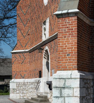 Zdjęcie nr 85: Kościół murowany z cegły, z użyciem zendrówki, na wysokim podmurowaniu z kamienia łupanego, jednonawowy, orientowany. 
Prezbiterium i nawa na planie prostokątów, nawa szersza i dłuższa od prezbiterium. Zakrystia na rzucie wydłużonego prostokąta przylega do wschodniej ściany prezbiterium, występując poza mury nawy. Prezbiterium od wschodu ujęte w narożach dwiema ukośnymi szkarpami, od południa jedną prostopadłą. Przy południowej ścianie nawy kruchta na planie kwadratu. Nawa w narożach ujęta szkarpami, od wschodu dwiema prostopadłymi, od zachodu ukośnymi. 
Prezbiterium dwuprzęsłowe, nakryte sklepieniem krzyżowo-żebrowym. Na zwornikach dwie tarcze z herbami Trzy Korony i Wieniawa. Zakrystia sklepiona kolebkowo. Nawa nakryta drewnianym stropem z kasetonami i rozetami. Kruchta sklepiona krzyżowo-żebrowo ze zwornikiem z tarczą z herbem Wieniawa. Otwór tęczowy w formie stojącego prostokąta zamkniętego łukiem ostrym. W zachodniej części nawy drewniany parapet chóru muzycznego wsparty na dwóch filarach. 
Fasada w jednej trzeciej wysokości odcięta kamiennym gzymsem kapnikowym, zakończona jodełkowym fryzem w partii muru, zwieńczona trójkątnym szczytem. Mur dekorowany zendrówką ułożoną „w zęby”, w partii szczytu w układzie rombowym. Na osi kamienny ostrołukowy portal o schodkowym nadprożu, dekorowanym krzyżującym się laskowaniem; węgary gładkie w jednej trzeciej wysokości przechodzące w piramidki i profilowanie powyżej. Nad portalem kamienna kwadratowa tablica, ujęta kamiennym obramieniem z tarczą z herbem Wieniawa. Ponad nią, na osi okulus w kamiennym obramieniu. 
Elewacje północna i południowa w części nawy w jednej trzeciej wysokości przedzielone kamiennym gzymsem kapnikowym, na zakończeniu ścian z gzymsem wieńczącym. Mur dekorowany zendrówką w układzie rombowym. Na elewacji południowej cztery okna w formie wydłużonego, stojącego prostokąta zamkniętego łukiem ostrym, w kamiennych obramieniach, podzielone laskowaniem. Południowa ściana prezbiterium zakończona gzymsem wieńczącym, w górnej części muru dekorowana zendrówką ułożoną „w zęby”. 
Szkarpy opinające nawę uskokowe, zwężające się ku górze, nakryte daszkami pulpitowymi. Szkarpy opinające prezbiterium nakryte daszkami pulpitowymi, podzielone gzymsami kapnikowymi. 
Kruchta od strony wschodniej i zachodniej podzielona kamiennym gzymsem kapnikowym, od południa zakończona gzymsem wieńczącym. Na ścianie południowej kruchty kamienny, ostrołukowy portal, o schodkowym nadprożu, dekorowanym krzyżującym się laskowaniem; węgary gładkie, w jednej trzeciej wysokości przechodzące w piramidki i profilowanie powyżej. Nad portalem kamienna tarcza z herbem Wieniawa. 
Elewacja wschodnia zwieńczona trójkątnym szczytem, w którego partii mur dekorowany jest zendrówką w układzie rombowym. Na osi okno w formie wydłużonego, stojącego prostokąta zamkniętego łukiem ostrym, w kamiennym obramieniu, zamurowane w trzech piątych wysokości, zakończone daszkiem pulpitowym. W dolnej części okna znajduje się ogrójec. 
Nawa i prezbiterium nakryte osobnymi blaszanymi dachami dwuspadowymi, kruchta i zakrystia pulpitowymi. Nad nawą blaszana wieżyczka na sygnaturkę z latarnią i hełmem baniastym.





