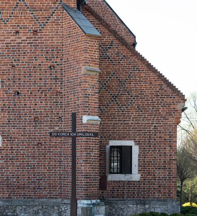 Zdjęcie nr 89: Kościół murowany z cegły, z użyciem zendrówki, na wysokim podmurowaniu z kamienia łupanego, jednonawowy, orientowany. 
Prezbiterium i nawa na planie prostokątów, nawa szersza i dłuższa od prezbiterium. Zakrystia na rzucie wydłużonego prostokąta przylega do wschodniej ściany prezbiterium, występując poza mury nawy. Prezbiterium od wschodu ujęte w narożach dwiema ukośnymi szkarpami, od południa jedną prostopadłą. Przy południowej ścianie nawy kruchta na planie kwadratu. Nawa w narożach ujęta szkarpami, od wschodu dwiema prostopadłymi, od zachodu ukośnymi. 
Prezbiterium dwuprzęsłowe, nakryte sklepieniem krzyżowo-żebrowym. Na zwornikach dwie tarcze z herbami Trzy Korony i Wieniawa. Zakrystia sklepiona kolebkowo. Nawa nakryta drewnianym stropem z kasetonami i rozetami. Kruchta sklepiona krzyżowo-żebrowo ze zwornikiem z tarczą z herbem Wieniawa. Otwór tęczowy w formie stojącego prostokąta zamkniętego łukiem ostrym. W zachodniej części nawy drewniany parapet chóru muzycznego wsparty na dwóch filarach. 
Fasada w jednej trzeciej wysokości odcięta kamiennym gzymsem kapnikowym, zakończona jodełkowym fryzem w partii muru, zwieńczona trójkątnym szczytem. Mur dekorowany zendrówką ułożoną „w zęby”, w partii szczytu w układzie rombowym. Na osi kamienny ostrołukowy portal o schodkowym nadprożu, dekorowanym krzyżującym się laskowaniem; węgary gładkie w jednej trzeciej wysokości przechodzące w piramidki i profilowanie powyżej. Nad portalem kamienna kwadratowa tablica, ujęta kamiennym obramieniem z tarczą z herbem Wieniawa. Ponad nią, na osi okulus w kamiennym obramieniu. 
Elewacje północna i południowa w części nawy w jednej trzeciej wysokości przedzielone kamiennym gzymsem kapnikowym, na zakończeniu ścian z gzymsem wieńczącym. Mur dekorowany zendrówką w układzie rombowym. Na elewacji południowej cztery okna w formie wydłużonego, stojącego prostokąta zamkniętego łukiem ostrym, w kamiennych obramieniach, podzielone laskowaniem. Południowa ściana prezbiterium zakończona gzymsem wieńczącym, w górnej części muru dekorowana zendrówką ułożoną „w zęby”. 
Szkarpy opinające nawę uskokowe, zwężające się ku górze, nakryte daszkami pulpitowymi. Szkarpy opinające prezbiterium nakryte daszkami pulpitowymi, podzielone gzymsami kapnikowymi. 
Kruchta od strony wschodniej i zachodniej podzielona kamiennym gzymsem kapnikowym, od południa zakończona gzymsem wieńczącym. Na ścianie południowej kruchty kamienny, ostrołukowy portal, o schodkowym nadprożu, dekorowanym krzyżującym się laskowaniem; węgary gładkie, w jednej trzeciej wysokości przechodzące w piramidki i profilowanie powyżej. Nad portalem kamienna tarcza z herbem Wieniawa. 
Elewacja wschodnia zwieńczona trójkątnym szczytem, w którego partii mur dekorowany jest zendrówką w układzie rombowym. Na osi okno w formie wydłużonego, stojącego prostokąta zamkniętego łukiem ostrym, w kamiennym obramieniu, zamurowane w trzech piątych wysokości, zakończone daszkiem pulpitowym. W dolnej części okna znajduje się ogrójec. 
Nawa i prezbiterium nakryte osobnymi blaszanymi dachami dwuspadowymi, kruchta i zakrystia pulpitowymi. Nad nawą blaszana wieżyczka na sygnaturkę z latarnią i hełmem baniastym.





