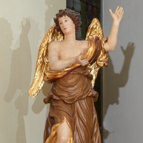 Zdjęcie nr 1: Rzeźba ścięta z tyłu, drążona. Figura anioła ustawiona na kłębiącym się obłoku, frontalnie, w lekkim kontrapoście, obie ręce uniesione ku górze, zwrócone w lewo, głowa lekko przechylona w prawo. Twarz pociągła, okolona ekspresyjnie rozwianymi włosami układającymi się w pojedyncze pukle. Anioł ubrany jest w sięgającą do ziemi szatę przewiązaną ukośnie przez lewe ramię, odsłaniającą fragment torsu oraz prawą nogę. Tkanina przylegająca do ciała jest silnie drapowana w liczne, drobne falujące fałdy, u spodu z wywiniętymi rozwianymi brzegami. Skrzydła niewielkie, złożone i złocone. Polichromia naturalistyczna, szata jasnobrązowa o złoconej podszewce, skrzydła złocone.