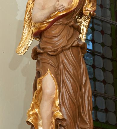 Zdjęcie nr 2: Rzeźba ścięta z tyłu, drążona. Figura anioła ustawiona na kłębiącym się obłoku, frontalnie, w lekkim kontrapoście, obie ręce uniesione ku górze, zwrócone w lewo, głowa lekko przechylona w prawo. Twarz pociągła, okolona ekspresyjnie rozwianymi włosami układającymi się w pojedyncze pukle. Anioł ubrany jest w sięgającą do ziemi szatę przewiązaną ukośnie przez lewe ramię, odsłaniającą fragment torsu oraz prawą nogę. Tkanina przylegająca do ciała jest silnie drapowana w liczne, drobne falujące fałdy, u spodu z wywiniętymi rozwianymi brzegami. Skrzydła niewielkie, złożone i złocone. Polichromia naturalistyczna, szata jasnobrązowa o złoconej podszewce, skrzydła złocone.