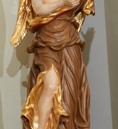 Zdjęcie nr 3: Rzeźba ścięta z tyłu, drążona. Figura anioła ustawiona na kłębiącym się obłoku, frontalnie, w lekkim kontrapoście, obie ręce uniesione ku górze, zwrócone w lewo, głowa lekko przechylona w prawo. Twarz pociągła, okolona ekspresyjnie rozwianymi włosami układającymi się w pojedyncze pukle. Anioł ubrany jest w sięgającą do ziemi szatę przewiązaną ukośnie przez lewe ramię, odsłaniającą fragment torsu oraz prawą nogę. Tkanina przylegająca do ciała jest silnie drapowana w liczne, drobne falujące fałdy, u spodu z wywiniętymi rozwianymi brzegami. Skrzydła niewielkie, złożone i złocone. Polichromia naturalistyczna, szata jasnobrązowa o złoconej podszewce, skrzydła złocone.