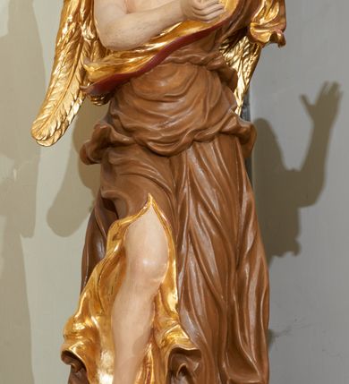 Zdjęcie nr 4: Rzeźba ścięta z tyłu, drążona. Figura anioła ustawiona na kłębiącym się obłoku, frontalnie, w lekkim kontrapoście, obie ręce uniesione ku górze, zwrócone w lewo, głowa lekko przechylona w prawo. Twarz pociągła, okolona ekspresyjnie rozwianymi włosami układającymi się w pojedyncze pukle. Anioł ubrany jest w sięgającą do ziemi szatę przewiązaną ukośnie przez lewe ramię, odsłaniającą fragment torsu oraz prawą nogę. Tkanina przylegająca do ciała jest silnie drapowana w liczne, drobne falujące fałdy, u spodu z wywiniętymi rozwianymi brzegami. Skrzydła niewielkie, złożone i złocone. Polichromia naturalistyczna, szata jasnobrązowa o złoconej podszewce, skrzydła złocone.