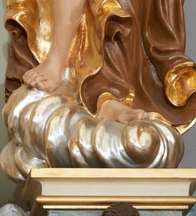 Zdjęcie nr 5: Rzeźba ścięta z tyłu, drążona. Figura anioła ustawiona na kłębiącym się obłoku, frontalnie, w lekkim kontrapoście, obie ręce uniesione ku górze, zwrócone w lewo, głowa lekko przechylona w prawo. Twarz pociągła, okolona ekspresyjnie rozwianymi włosami układającymi się w pojedyncze pukle. Anioł ubrany jest w sięgającą do ziemi szatę przewiązaną ukośnie przez lewe ramię, odsłaniającą fragment torsu oraz prawą nogę. Tkanina przylegająca do ciała jest silnie drapowana w liczne, drobne falujące fałdy, u spodu z wywiniętymi rozwianymi brzegami. Skrzydła niewielkie, złożone i złocone. Polichromia naturalistyczna, szata jasnobrązowa o złoconej podszewce, skrzydła złocone.