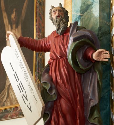 Zdjęcie nr 1: Rzeźba przedstawiająca proroka Mojżesza, ścięta z tyłu, drążona. Prorok ustawiony na niskim cokole w formie posrebrzonego obłoku. Ukazany jest frontalnie w delikatnym kontrapoście z lewą nogą ugiętą w kolanie i z rękami wyciągniętymi szeroko w boki, z głową skierowaną w lewo i uniesioną do góry. W prawej dłoni trzyma tablice Dekalogu. Twarz podłużna z długim nosem, otwartymi ustami i zmarszczonymi łukami brwiowymi; okolona długą, ciemną brodą i średniej długości włosami, zasłaniającymi uszy i uformowanymi nad czołem w dwa rogi. Ubrany jest w długą, czerwoną suknię z kołnierzem, przewiązaną w talii, obficie drapowaną oraz zielony płaszcz z fioletową podszewką, zawinięty wokół lewego ramienia; na stopach ma założone pełne, brązowe buty. Polichromia w odsłoniętych partiach ciała jest naturalistyczna, policzki zaczerwienione.



