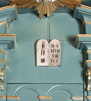 Zdjęcie nr 3: Ambona przyścienna, wisząca, z dostępem od zakrystii. Kosz na planie czterech boków z sześcioboku, na cokole w formie spłaszczonego wałka, z podwieszonym niewielkim cokolikiem zakończonym szyszką. Ścianki wydzielone pilastrami o kanelowanych trzonach, na cokołach, dźwigającymi parapet w formie gzymsu, wyłamany na ich osi. W polach ścianek płyciny o kształcie stojącego prostokąta z płaskorzeźbionymi przedstawieniami czterech ewangelistów. Zaplecek w formie wydłużonego, stojącego prostokąta, ujętego pilastrami o kanelowanych trzonach, zakończonych konsolami dekorowanymi liściem akantu, dźwigającymi imposty oraz przerwany, półokrągły przyczółek. W polu zaplecka drzwiczki do zakrystii, zdobione dwiema arkadami; ponad nimi płaskorzeźbione tablice praw. Baldachim na planie analogicznym względem kosza, w formie kopułki na wklęsłym, zwężającym się cokole dekorowanym płycinami, zakończonej latarnią z krzyżem na kuli. Brzeg w postaci gzymsu wyłamanego na osi podpór kosza, z siedzącymi nań puttami oraz z podwieszonym lambrekinem. Na podniebiu baldachimu rzeźbiona gołębica Ducha Świętego w glorii promienistej. 
Struktura polichromowana w kolorze szaro-niebieskim, profile i ornamentyka złocone.