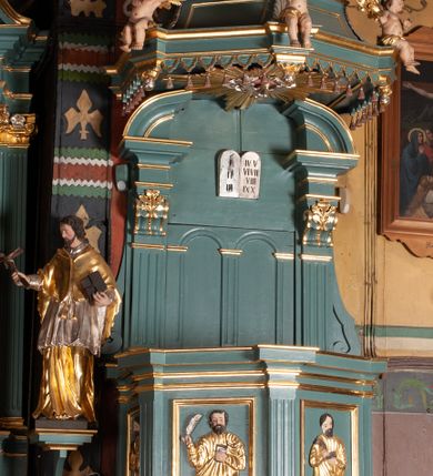 Zdjęcie nr 2: Ambona przyścienna, wisząca, z dostępem od zakrystii. Kosz na planie czterech boków z sześcioboku, na cokole w formie spłaszczonego wałka, z podwieszonym niewielkim cokolikiem zakończonym szyszką. Ścianki wydzielone pilastrami o kanelowanych trzonach, na cokołach, dźwigającymi parapet w formie gzymsu, wyłamany na ich osi. W polach ścianek płyciny o kształcie stojącego prostokąta z płaskorzeźbionymi przedstawieniami czterech ewangelistów. Zaplecek w formie wydłużonego, stojącego prostokąta, ujętego pilastrami o kanelowanych trzonach, zakończonych konsolami dekorowanymi liściem akantu, dźwigającymi imposty oraz przerwany, półokrągły przyczółek. W polu zaplecka drzwiczki do zakrystii, zdobione dwiema arkadami; ponad nimi płaskorzeźbione tablice praw. Baldachim na planie analogicznym względem kosza, w formie kopułki na wklęsłym, zwężającym się cokole dekorowanym płycinami, zakończonej latarnią z krzyżem na kuli. Brzeg w postaci gzymsu wyłamanego na osi podpór kosza, z siedzącymi nań puttami oraz z podwieszonym lambrekinem. Na podniebiu baldachimu rzeźbiona gołębica Ducha Świętego w glorii promienistej. 
Struktura polichromowana w kolorze szaro-niebieskim, profile i ornamentyka złocone.