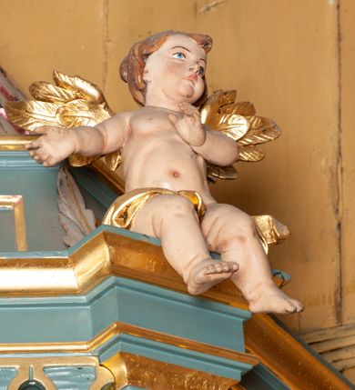 Zdjęcie nr 12: Ambona przyścienna, wisząca, z dostępem od zakrystii. Kosz na planie czterech boków z sześcioboku, na cokole w formie spłaszczonego wałka, z podwieszonym niewielkim cokolikiem zakończonym szyszką. Ścianki wydzielone pilastrami o kanelowanych trzonach, na cokołach, dźwigającymi parapet w formie gzymsu, wyłamany na ich osi. W polach ścianek płyciny o kształcie stojącego prostokąta z płaskorzeźbionymi przedstawieniami czterech ewangelistów. Zaplecek w formie wydłużonego, stojącego prostokąta, ujętego pilastrami o kanelowanych trzonach, zakończonych konsolami dekorowanymi liściem akantu, dźwigającymi imposty oraz przerwany, półokrągły przyczółek. W polu zaplecka drzwiczki do zakrystii, zdobione dwiema arkadami; ponad nimi płaskorzeźbione tablice praw. Baldachim na planie analogicznym względem kosza, w formie kopułki na wklęsłym, zwężającym się cokole dekorowanym płycinami, zakończonej latarnią z krzyżem na kuli. Brzeg w postaci gzymsu wyłamanego na osi podpór kosza, z siedzącymi nań puttami oraz z podwieszonym lambrekinem. Na podniebiu baldachimu rzeźbiona gołębica Ducha Świętego w glorii promienistej. 
Struktura polichromowana w kolorze szaro-niebieskim, profile i ornamentyka złocone.
