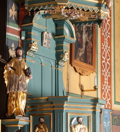 Zdjęcie nr 1: Ambona przyścienna, wisząca, z dostępem od zakrystii. Kosz na planie czterech boków z sześcioboku, na cokole w formie spłaszczonego wałka, z podwieszonym niewielkim cokolikiem zakończonym szyszką. Ścianki wydzielone pilastrami o kanelowanych trzonach, na cokołach, dźwigającymi parapet w formie gzymsu, wyłamany na ich osi. W polach ścianek płyciny o kształcie stojącego prostokąta z płaskorzeźbionymi przedstawieniami czterech ewangelistów. Zaplecek w formie wydłużonego, stojącego prostokąta, ujętego pilastrami o kanelowanych trzonach, zakończonych konsolami dekorowanymi liściem akantu, dźwigającymi imposty oraz przerwany, półokrągły przyczółek. W polu zaplecka drzwiczki do zakrystii, zdobione dwiema arkadami; ponad nimi płaskorzeźbione tablice praw. Baldachim na planie analogicznym względem kosza, w formie kopułki na wklęsłym, zwężającym się cokole dekorowanym płycinami, zakończonej latarnią z krzyżem na kuli. Brzeg w postaci gzymsu wyłamanego na osi podpór kosza, z siedzącymi nań puttami oraz z podwieszonym lambrekinem. Na podniebiu baldachimu rzeźbiona gołębica Ducha Świętego w glorii promienistej. 
Struktura polichromowana w kolorze szaro-niebieskim, profile i ornamentyka złocone.