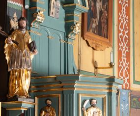 Zdjęcie nr 1: Ambona przyścienna, wisząca, z dostępem od zakrystii. Kosz na planie czterech boków z sześcioboku, na cokole w formie spłaszczonego wałka, z podwieszonym niewielkim cokolikiem zakończonym szyszką. Ścianki wydzielone pilastrami o kanelowanych trzonach, na cokołach, dźwigającymi parapet w formie gzymsu, wyłamany na ich osi. W polach ścianek płyciny o kształcie stojącego prostokąta z płaskorzeźbionymi przedstawieniami czterech ewangelistów. Zaplecek w formie wydłużonego, stojącego prostokąta, ujętego pilastrami o kanelowanych trzonach, zakończonych konsolami dekorowanymi liściem akantu, dźwigającymi imposty oraz przerwany, półokrągły przyczółek. W polu zaplecka drzwiczki do zakrystii, zdobione dwiema arkadami; ponad nimi płaskorzeźbione tablice praw. Baldachim na planie analogicznym względem kosza, w formie kopułki na wklęsłym, zwężającym się cokole dekorowanym płycinami, zakończonej latarnią z krzyżem na kuli. Brzeg w postaci gzymsu wyłamanego na osi podpór kosza, z siedzącymi nań puttami oraz z podwieszonym lambrekinem. Na podniebiu baldachimu rzeźbiona gołębica Ducha Świętego w glorii promienistej. 
Struktura polichromowana w kolorze szaro-niebieskim, profile i ornamentyka złocone.