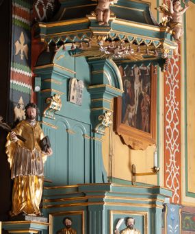 Zdjęcie nr 1: Ambona przyścienna, wisząca, z dostępem od zakrystii. Kosz na planie czterech boków z sześcioboku, na cokole w formie spłaszczonego wałka, z podwieszonym niewielkim cokolikiem zakończonym szyszką. Ścianki wydzielone pilastrami o kanelowanych trzonach, na cokołach, dźwigającymi parapet w formie gzymsu, wyłamany na ich osi. W polach ścianek płyciny o kształcie stojącego prostokąta z płaskorzeźbionymi przedstawieniami czterech ewangelistów. Zaplecek w formie wydłużonego, stojącego prostokąta, ujętego pilastrami o kanelowanych trzonach, zakończonych konsolami dekorowanymi liściem akantu, dźwigającymi imposty oraz przerwany, półokrągły przyczółek. W polu zaplecka drzwiczki do zakrystii, zdobione dwiema arkadami; ponad nimi płaskorzeźbione tablice praw. Baldachim na planie analogicznym względem kosza, w formie kopułki na wklęsłym, zwężającym się cokole dekorowanym płycinami, zakończonej latarnią z krzyżem na kuli. Brzeg w postaci gzymsu wyłamanego na osi podpór kosza, z siedzącymi nań puttami oraz z podwieszonym lambrekinem. Na podniebiu baldachimu rzeźbiona gołębica Ducha Świętego w glorii promienistej. 
Struktura polichromowana w kolorze szaro-niebieskim, profile i ornamentyka złocone.