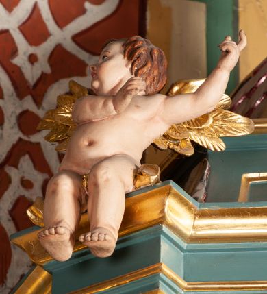 Zdjęcie nr 10: Ambona przyścienna, wisząca, z dostępem od zakrystii. Kosz na planie czterech boków z sześcioboku, na cokole w formie spłaszczonego wałka, z podwieszonym niewielkim cokolikiem zakończonym szyszką. Ścianki wydzielone pilastrami o kanelowanych trzonach, na cokołach, dźwigającymi parapet w formie gzymsu, wyłamany na ich osi. W polach ścianek płyciny o kształcie stojącego prostokąta z płaskorzeźbionymi przedstawieniami czterech ewangelistów. Zaplecek w formie wydłużonego, stojącego prostokąta, ujętego pilastrami o kanelowanych trzonach, zakończonych konsolami dekorowanymi liściem akantu, dźwigającymi imposty oraz przerwany, półokrągły przyczółek. W polu zaplecka drzwiczki do zakrystii, zdobione dwiema arkadami; ponad nimi płaskorzeźbione tablice praw. Baldachim na planie analogicznym względem kosza, w formie kopułki na wklęsłym, zwężającym się cokole dekorowanym płycinami, zakończonej latarnią z krzyżem na kuli. Brzeg w postaci gzymsu wyłamanego na osi podpór kosza, z siedzącymi nań puttami oraz z podwieszonym lambrekinem. Na podniebiu baldachimu rzeźbiona gołębica Ducha Świętego w glorii promienistej. 
Struktura polichromowana w kolorze szaro-niebieskim, profile i ornamentyka złocone.