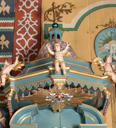 Zdjęcie nr 5: Ambona przyścienna, wisząca, z dostępem od zakrystii. Kosz na planie czterech boków z sześcioboku, na cokole w formie spłaszczonego wałka, z podwieszonym niewielkim cokolikiem zakończonym szyszką. Ścianki wydzielone pilastrami o kanelowanych trzonach, na cokołach, dźwigającymi parapet w formie gzymsu, wyłamany na ich osi. W polach ścianek płyciny o kształcie stojącego prostokąta z płaskorzeźbionymi przedstawieniami czterech ewangelistów. Zaplecek w formie wydłużonego, stojącego prostokąta, ujętego pilastrami o kanelowanych trzonach, zakończonych konsolami dekorowanymi liściem akantu, dźwigającymi imposty oraz przerwany, półokrągły przyczółek. W polu zaplecka drzwiczki do zakrystii, zdobione dwiema arkadami; ponad nimi płaskorzeźbione tablice praw. Baldachim na planie analogicznym względem kosza, w formie kopułki na wklęsłym, zwężającym się cokole dekorowanym płycinami, zakończonej latarnią z krzyżem na kuli. Brzeg w postaci gzymsu wyłamanego na osi podpór kosza, z siedzącymi nań puttami oraz z podwieszonym lambrekinem. Na podniebiu baldachimu rzeźbiona gołębica Ducha Świętego w glorii promienistej. 
Struktura polichromowana w kolorze szaro-niebieskim, profile i ornamentyka złocone.