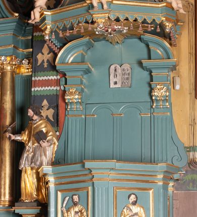 Zdjęcie nr 4: Ambona przyścienna, wisząca, z dostępem od zakrystii. Kosz na planie czterech boków z sześcioboku, na cokole w formie spłaszczonego wałka, z podwieszonym niewielkim cokolikiem zakończonym szyszką. Ścianki wydzielone pilastrami o kanelowanych trzonach, na cokołach, dźwigającymi parapet w formie gzymsu, wyłamany na ich osi. W polach ścianek płyciny o kształcie stojącego prostokąta z płaskorzeźbionymi przedstawieniami czterech ewangelistów. Zaplecek w formie wydłużonego, stojącego prostokąta, ujętego pilastrami o kanelowanych trzonach, zakończonych konsolami dekorowanymi liściem akantu, dźwigającymi imposty oraz przerwany, półokrągły przyczółek. W polu zaplecka drzwiczki do zakrystii, zdobione dwiema arkadami; ponad nimi płaskorzeźbione tablice praw. Baldachim na planie analogicznym względem kosza, w formie kopułki na wklęsłym, zwężającym się cokole dekorowanym płycinami, zakończonej latarnią z krzyżem na kuli. Brzeg w postaci gzymsu wyłamanego na osi podpór kosza, z siedzącymi nań puttami oraz z podwieszonym lambrekinem. Na podniebiu baldachimu rzeźbiona gołębica Ducha Świętego w glorii promienistej. 
Struktura polichromowana w kolorze szaro-niebieskim, profile i ornamentyka złocone.