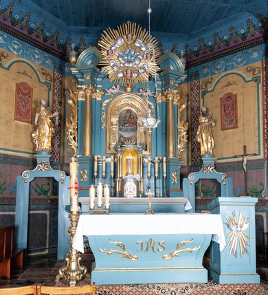 Zdjęcie nr 3: Ołtarz architektoniczny, jednokondygnacyjny, jednoosiowy, z bramkami i zwieńczeniem. Ołtarz kamienny, sarkofagowy, wklęsło-wypukły, na cokole. Mensa kamienna, o brzegu w formie profilowanego gzymsu. Tabernakulum drewniane, obite blachą, w postaci prostopadłościennej skrzynki, z wypukłą ścianką frontową, flankowane parą drewnianych kolumienek o kapitelach kompozytowych, na cokołach. Drzwiczki w formie kwadratu zamkniętego łukiem półkolistym, ujęte ramą z odlewaną dekoracją roślinną, w polu aplikowane, odlewane przedstawienie kielicha eucharystycznego w glorii promienistej na obłoku, z kłosami zbóż po bokach. Na tabernakulum osadzony tron wystawienia w formie prostopadłościanu, z wypukłą ścianką frontową oraz obrotową niszą ujętą profilowaną ramą, flankowany parą kolumienek o kapitelach kompozytowych, dźwigających imposty oraz przerwane w partii architrawu i fryzu belkowanie opasujące go z trzech stron. Na osi podpór przerwany, segmentowy przyczółek, ustawiony pod kątem. Na belkowaniu figurka Baranka Apokaliptycznego, leżącego na księdze na tle glorii promienistej. 
Retabulum na dwustrefowym cokole, w górnej partii zryzalitowanym przy brzegach, ujęte parą zwielokrotnionych pilastrów oraz stojącą przed nimi parą kolumn o kapitelach kompozytowych, ustawionych pod katem względem lica. Podpory dźwigają pełne belkowanie, wyłamane na ich osi, przerwane nad polem środkowym; na linii kolumn przerwany półokrągły przyczółek. W polu zwieńczenia gloria promienista z rzeźbioną gołębicą Ducha Świętego w otoczeniu obłoków i uskrzydlonych główek anielskich. W polu środkowym nisza w formie stojącego prostokąta zamkniętego łukiem segmentowym, w profilowanej ramie, z obrazem Matki Boskiej z Dzieciątkiem w aediculi. Bramki z przejściami w formie wydłużonego, stojącego prostokąta o wklęsłych górnych narożach, zamknięte dwoma spływami wolutowymi stanowiącymi podstawę dla cokołów na figury św. Piotra i św. Pawła. 
Cokoły kolumn dekorowane akantem. W polu środkowym, nad niszą para puttów podtrzymujących koronę; ramę flankują rocaillowe zwisy; u jej spodu palmety i rozety. Na brzegach belkowania wazony dekorowane plecionką i liśćmi. Kondygnacja ujęta uszami utworzonymi z wolut i liści palmowych. Spód cokołu wieńczącego bramki dekorowany muszlą; woluty zdobione kampanulami.
 Struktura polichromowana w kolorze szaro-niebieskim; kolumny, ramy, gloria, profile i ornamentyka złocone; obłoki srebrzone.
