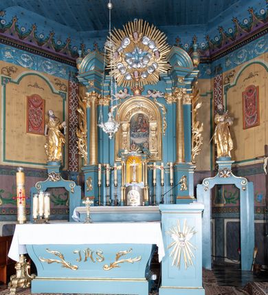 Zdjęcie nr 2: Ołtarz architektoniczny, jednokondygnacyjny, jednoosiowy, z bramkami i zwieńczeniem. Ołtarz kamienny, sarkofagowy, wklęsło-wypukły, na cokole. Mensa kamienna, o brzegu w formie profilowanego gzymsu. Tabernakulum drewniane, obite blachą, w postaci prostopadłościennej skrzynki, z wypukłą ścianką frontową, flankowane parą drewnianych kolumienek o kapitelach kompozytowych, na cokołach. Drzwiczki w formie kwadratu zamkniętego łukiem półkolistym, ujęte ramą z odlewaną dekoracją roślinną, w polu aplikowane, odlewane przedstawienie kielicha eucharystycznego w glorii promienistej na obłoku, z kłosami zbóż po bokach. Na tabernakulum osadzony tron wystawienia w formie prostopadłościanu, z wypukłą ścianką frontową oraz obrotową niszą ujętą profilowaną ramą, flankowany parą kolumienek o kapitelach kompozytowych, dźwigających imposty oraz przerwane w partii architrawu i fryzu belkowanie opasujące go z trzech stron. Na osi podpór przerwany, segmentowy przyczółek, ustawiony pod kątem. Na belkowaniu figurka Baranka Apokaliptycznego, leżącego na księdze na tle glorii promienistej. 
Retabulum na dwustrefowym cokole, w górnej partii zryzalitowanym przy brzegach, ujęte parą zwielokrotnionych pilastrów oraz stojącą przed nimi parą kolumn o kapitelach kompozytowych, ustawionych pod katem względem lica. Podpory dźwigają pełne belkowanie, wyłamane na ich osi, przerwane nad polem środkowym; na linii kolumn przerwany półokrągły przyczółek. W polu zwieńczenia gloria promienista z rzeźbioną gołębicą Ducha Świętego w otoczeniu obłoków i uskrzydlonych główek anielskich. W polu środkowym nisza w formie stojącego prostokąta zamkniętego łukiem segmentowym, w profilowanej ramie, z obrazem Matki Boskiej z Dzieciątkiem w aediculi. Bramki z przejściami w formie wydłużonego, stojącego prostokąta o wklęsłych górnych narożach, zamknięte dwoma spływami wolutowymi stanowiącymi podstawę dla cokołów na figury św. Piotra i św. Pawła. 
Cokoły kolumn dekorowane akantem. W polu środkowym, nad niszą para puttów podtrzymujących koronę; ramę flankują rocaillowe zwisy; u jej spodu palmety i rozety. Na brzegach belkowania wazony dekorowane plecionką i liśćmi. Kondygnacja ujęta uszami utworzonymi z wolut i liści palmowych. Spód cokołu wieńczącego bramki dekorowany muszlą; woluty zdobione kampanulami.
 Struktura polichromowana w kolorze szaro-niebieskim; kolumny, ramy, gloria, profile i ornamentyka złocone; obłoki srebrzone.

