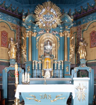 Zdjęcie nr 1: Ołtarz architektoniczny, jednokondygnacyjny, jednoosiowy, z bramkami i zwieńczeniem. Ołtarz kamienny, sarkofagowy, wklęsło-wypukły, na cokole. Mensa kamienna, o brzegu w formie profilowanego gzymsu. Tabernakulum drewniane, obite blachą, w postaci prostopadłościennej skrzynki, z wypukłą ścianką frontową, flankowane parą drewnianych kolumienek o kapitelach kompozytowych, na cokołach. Drzwiczki w formie kwadratu zamkniętego łukiem półkolistym, ujęte ramą z odlewaną dekoracją roślinną, w polu aplikowane, odlewane przedstawienie kielicha eucharystycznego w glorii promienistej na obłoku, z kłosami zbóż po bokach. Na tabernakulum osadzony tron wystawienia w formie prostopadłościanu, z wypukłą ścianką frontową oraz obrotową niszą ujętą profilowaną ramą, flankowany parą kolumienek o kapitelach kompozytowych, dźwigających imposty oraz przerwane w partii architrawu i fryzu belkowanie opasujące go z trzech stron. Na osi podpór przerwany, segmentowy przyczółek, ustawiony pod kątem. Na belkowaniu figurka Baranka Apokaliptycznego, leżącego na księdze na tle glorii promienistej. 
Retabulum na dwustrefowym cokole, w górnej partii zryzalitowanym przy brzegach, ujęte parą zwielokrotnionych pilastrów oraz stojącą przed nimi parą kolumn o kapitelach kompozytowych, ustawionych pod katem względem lica. Podpory dźwigają pełne belkowanie, wyłamane na ich osi, przerwane nad polem środkowym; na linii kolumn przerwany półokrągły przyczółek. W polu zwieńczenia gloria promienista z rzeźbioną gołębicą Ducha Świętego w otoczeniu obłoków i uskrzydlonych główek anielskich. W polu środkowym nisza w formie stojącego prostokąta zamkniętego łukiem segmentowym, w profilowanej ramie, z obrazem Matki Boskiej z Dzieciątkiem w aediculi. Bramki z przejściami w formie wydłużonego, stojącego prostokąta o wklęsłych górnych narożach, zamknięte dwoma spływami wolutowymi stanowiącymi podstawę dla cokołów na figury św. Piotra i św. Pawła. 
Cokoły kolumn dekorowane akantem. W polu środkowym, nad niszą para puttów podtrzymujących koronę; ramę flankują rocaillowe zwisy; u jej spodu palmety i rozety. Na brzegach belkowania wazony dekorowane plecionką i liśćmi. Kondygnacja ujęta uszami utworzonymi z wolut i liści palmowych. Spód cokołu wieńczącego bramki dekorowany muszlą; woluty zdobione kampanulami.
 Struktura polichromowana w kolorze szaro-niebieskim; kolumny, ramy, gloria, profile i ornamentyka złocone; obłoki srebrzone.
