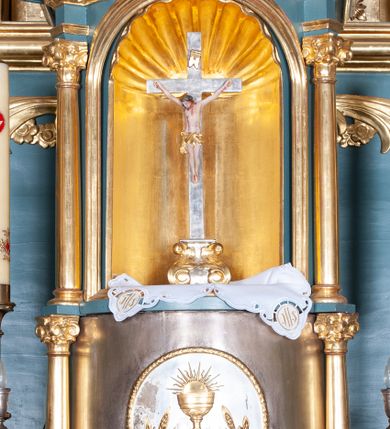 Zdjęcie nr 8: Ołtarz architektoniczny, jednokondygnacyjny, jednoosiowy, z bramkami i zwieńczeniem. Ołtarz kamienny, sarkofagowy, wklęsło-wypukły, na cokole. Mensa kamienna, o brzegu w formie profilowanego gzymsu. Tabernakulum drewniane, obite blachą, w postaci prostopadłościennej skrzynki, z wypukłą ścianką frontową, flankowane parą drewnianych kolumienek o kapitelach kompozytowych, na cokołach. Drzwiczki w formie kwadratu zamkniętego łukiem półkolistym, ujęte ramą z odlewaną dekoracją roślinną, w polu aplikowane, odlewane przedstawienie kielicha eucharystycznego w glorii promienistej na obłoku, z kłosami zbóż po bokach. Na tabernakulum osadzony tron wystawienia w formie prostopadłościanu, z wypukłą ścianką frontową oraz obrotową niszą ujętą profilowaną ramą, flankowany parą kolumienek o kapitelach kompozytowych, dźwigających imposty oraz przerwane w partii architrawu i fryzu belkowanie opasujące go z trzech stron. Na osi podpór przerwany, segmentowy przyczółek, ustawiony pod kątem. Na belkowaniu figurka Baranka Apokaliptycznego, leżącego na księdze na tle glorii promienistej. 
Retabulum na dwustrefowym cokole, w górnej partii zryzalitowanym przy brzegach, ujęte parą zwielokrotnionych pilastrów oraz stojącą przed nimi parą kolumn o kapitelach kompozytowych, ustawionych pod katem względem lica. Podpory dźwigają pełne belkowanie, wyłamane na ich osi, przerwane nad polem środkowym; na linii kolumn przerwany półokrągły przyczółek. W polu zwieńczenia gloria promienista z rzeźbioną gołębicą Ducha Świętego w otoczeniu obłoków i uskrzydlonych główek anielskich. W polu środkowym nisza w formie stojącego prostokąta zamkniętego łukiem segmentowym, w profilowanej ramie, z obrazem Matki Boskiej z Dzieciątkiem w aediculi. Bramki z przejściami w formie wydłużonego, stojącego prostokąta o wklęsłych górnych narożach, zamknięte dwoma spływami wolutowymi stanowiącymi podstawę dla cokołów na figury św. Piotra i św. Pawła. 
Cokoły kolumn dekorowane akantem. W polu środkowym, nad niszą para puttów podtrzymujących koronę; ramę flankują rocaillowe zwisy; u jej spodu palmety i rozety. Na brzegach belkowania wazony dekorowane plecionką i liśćmi. Kondygnacja ujęta uszami utworzonymi z wolut i liści palmowych. Spód cokołu wieńczącego bramki dekorowany muszlą; woluty zdobione kampanulami.
 Struktura polichromowana w kolorze szaro-niebieskim; kolumny, ramy, gloria, profile i ornamentyka złocone; obłoki srebrzone.
