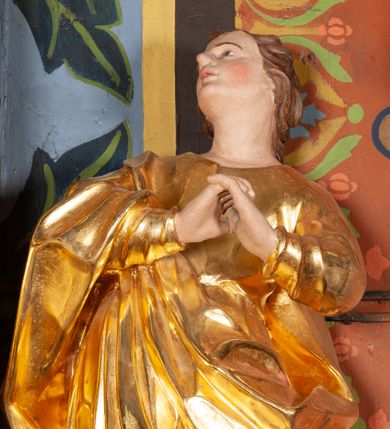 Zdjęcie nr 1: Całopostaciowa, lekko ścięta z tyłu figura św. Jana Ewangelisty zwróconej na wprost i stojącej w wydatnym kontrapoście. Sylwetka ciała esowato wygięta, tułów i głowa odchylone do tyłu. Głowa uniesiona, zwrócona w prawą stronę. Twarz o łagodnych rysach, okolona lekko falowanymi włosami. Czoło wydatne z pojedynczym lokiem włosów. Oczy niewielkie, usta rozchylone. Ręce uniesione na wysokość klatki piersiowej złożone w geście modlitewnym. Postać ubrana w długą tunikę oraz płaszcz, przerzucony przez prawe ramię, okrywający dolną część postaci. Karnacja ciała w jasnokremowym odcieniu, szaty złocone.

