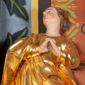 Zdjęcie nr 1: Całopostaciowa, lekko ścięta z tyłu figura św. Jana Ewangelisty zwróconej na wprost i stojącej w wydatnym kontrapoście. Sylwetka ciała esowato wygięta, tułów i głowa odchylone do tyłu. Głowa uniesiona, zwrócona w prawą stronę. Twarz o łagodnych rysach, okolona lekko falowanymi włosami. Czoło wydatne z pojedynczym lokiem włosów. Oczy niewielkie, usta rozchylone. Ręce uniesione na wysokość klatki piersiowej złożone w geście modlitewnym. Postać ubrana w długą tunikę oraz płaszcz, przerzucony przez prawe ramię, okrywający dolną część postaci. Karnacja ciała w jasnokremowym odcieniu, szaty złocone.

