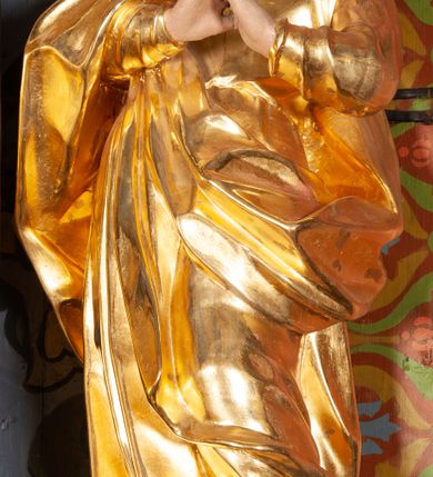 Zdjęcie nr 2: Całopostaciowa, lekko ścięta z tyłu figura św. Jana Ewangelisty zwróconej na wprost i stojącej w wydatnym kontrapoście. Sylwetka ciała esowato wygięta, tułów i głowa odchylone do tyłu. Głowa uniesiona, zwrócona w prawą stronę. Twarz o łagodnych rysach, okolona lekko falowanymi włosami. Czoło wydatne z pojedynczym lokiem włosów. Oczy niewielkie, usta rozchylone. Ręce uniesione na wysokość klatki piersiowej złożone w geście modlitewnym. Postać ubrana w długą tunikę oraz płaszcz, przerzucony przez prawe ramię, okrywający dolną część postaci. Karnacja ciała w jasnokremowym odcieniu, szaty złocone.

