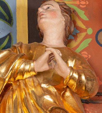 Zdjęcie nr 3: Całopostaciowa, lekko ścięta z tyłu figura św. Jana Ewangelisty zwróconej na wprost i stojącej w wydatnym kontrapoście. Sylwetka ciała esowato wygięta, tułów i głowa odchylone do tyłu. Głowa uniesiona, zwrócona w prawą stronę. Twarz o łagodnych rysach, okolona lekko falowanymi włosami. Czoło wydatne z pojedynczym lokiem włosów. Oczy niewielkie, usta rozchylone. Ręce uniesione na wysokość klatki piersiowej złożone w geście modlitewnym. Postać ubrana w długą tunikę oraz płaszcz, przerzucony przez prawe ramię, okrywający dolną część postaci. Karnacja ciała w jasnokremowym odcieniu, szaty złocone.

