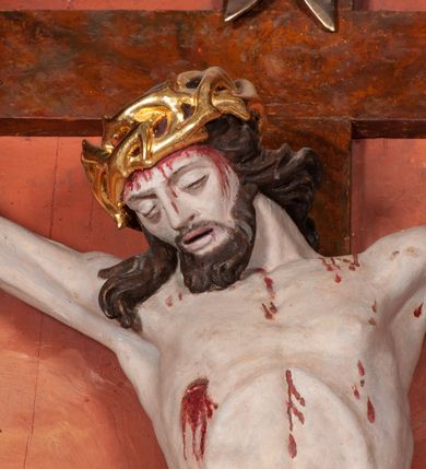 Zdjęcie nr 3: Figura Chrystusa ścięta z tyłu, przybita do belki krzyża trzema gwoździami, w typie Christo morte. Ciało w głębokim zwisie, ramiona ułożone w kształt litery „Y”. Głowa opadająca na klatkę piersiową, przechylona na prawe ramię. Twarz szeroka, okolona krótką brodą i włosami opadającymi na prawe ramię. Oczy zamknięte, nos niewielki, prosty, usta rozchylone. Na głowie korona cierniowa. Sylwetka ciała smukła o wyraźnie zaznaczonej muskulaturze, zwłaszcza w partii klatki piersiowej i ramion. Dłonie o zaciśniętych palcach. Wokół bioder perizonium układające się w głębokie poprzeczne fałdy, z rozwianym zwisem przy lewym boku. Stopy skrzyżowane, prawa założona na lewą. Karnacja ciała w jasnoszarym odcieniu z zaznaczonymi czerwoną farbą śladami męki. Włosy i zarost ciemnobrązowe, perizonium i korona cierniowa złote. Nad głową srebrny titulus w formie banderoli.