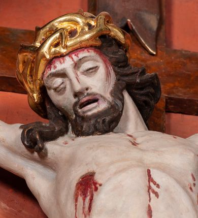 Zdjęcie nr 5: Figura Chrystusa ścięta z tyłu, przybita do belki krzyża trzema gwoździami, w typie Christo morte. Ciało w głębokim zwisie, ramiona ułożone w kształt litery „Y”. Głowa opadająca na klatkę piersiową, przechylona na prawe ramię. Twarz szeroka, okolona krótką brodą i włosami opadającymi na prawe ramię. Oczy zamknięte, nos niewielki, prosty, usta rozchylone. Na głowie korona cierniowa. Sylwetka ciała smukła o wyraźnie zaznaczonej muskulaturze, zwłaszcza w partii klatki piersiowej i ramion. Dłonie o zaciśniętych palcach. Wokół bioder perizonium układające się w głębokie poprzeczne fałdy, z rozwianym zwisem przy lewym boku. Stopy skrzyżowane, prawa założona na lewą. Karnacja ciała w jasnoszarym odcieniu z zaznaczonymi czerwoną farbą śladami męki. Włosy i zarost ciemnobrązowe, perizonium i korona cierniowa złote. Nad głową srebrny titulus w formie banderoli.