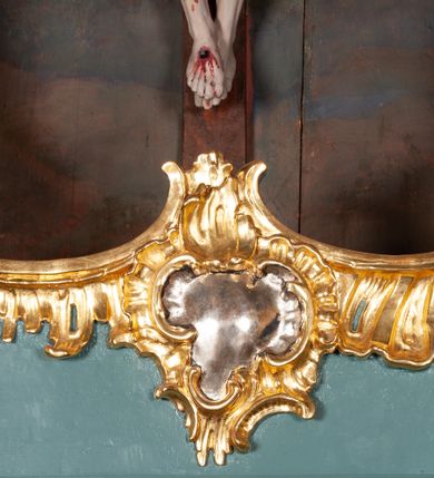 Zdjęcie nr 6: Figura Chrystusa ścięta z tyłu, przybita do belki krzyża trzema gwoździami, w typie Christo morte. Ciało w głębokim zwisie, ramiona ułożone w kształt litery „Y”. Głowa opadająca na klatkę piersiową, przechylona na prawe ramię. Twarz szeroka, okolona krótką brodą i włosami opadającymi na prawe ramię. Oczy zamknięte, nos niewielki, prosty, usta rozchylone. Na głowie korona cierniowa. Sylwetka ciała smukła o wyraźnie zaznaczonej muskulaturze, zwłaszcza w partii klatki piersiowej i ramion. Dłonie o zaciśniętych palcach. Wokół bioder perizonium układające się w głębokie poprzeczne fałdy, z rozwianym zwisem przy lewym boku. Stopy skrzyżowane, prawa założona na lewą. Karnacja ciała w jasnoszarym odcieniu z zaznaczonymi czerwoną farbą śladami męki. Włosy i zarost ciemnobrązowe, perizonium i korona cierniowa złote. Nad głową srebrny titulus w formie banderoli.