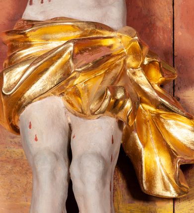 Zdjęcie nr 4: Figura Chrystusa ścięta z tyłu, przybita do belki krzyża trzema gwoździami, w typie Christo morte. Ciało w głębokim zwisie, ramiona ułożone w kształt litery „Y”. Głowa opadająca na klatkę piersiową, przechylona na prawe ramię. Twarz szeroka, okolona krótką brodą i włosami opadającymi na prawe ramię. Oczy zamknięte, nos niewielki, prosty, usta rozchylone. Na głowie korona cierniowa. Sylwetka ciała smukła o wyraźnie zaznaczonej muskulaturze, zwłaszcza w partii klatki piersiowej i ramion. Dłonie o zaciśniętych palcach. Wokół bioder perizonium układające się w głębokie poprzeczne fałdy, z rozwianym zwisem przy lewym boku. Stopy skrzyżowane, prawa założona na lewą. Karnacja ciała w jasnoszarym odcieniu z zaznaczonymi czerwoną farbą śladami męki. Włosy i zarost ciemnobrązowe, perizonium i korona cierniowa złote. Nad głową srebrny titulus w formie banderoli.