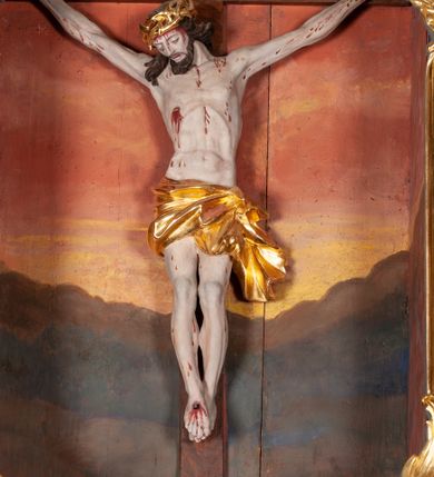 Zdjęcie nr 1: Figura Chrystusa ścięta z tyłu, przybita do belki krzyża trzema gwoździami, w typie Christo morte. Ciało w głębokim zwisie, ramiona ułożone w kształt litery „Y”. Głowa opadająca na klatkę piersiową, przechylona na prawe ramię. Twarz szeroka, okolona krótką brodą i włosami opadającymi na prawe ramię. Oczy zamknięte, nos niewielki, prosty, usta rozchylone. Na głowie korona cierniowa. Sylwetka ciała smukła o wyraźnie zaznaczonej muskulaturze, zwłaszcza w partii klatki piersiowej i ramion. Dłonie o zaciśniętych palcach. Wokół bioder perizonium układające się w głębokie poprzeczne fałdy, z rozwianym zwisem przy lewym boku. Stopy skrzyżowane, prawa założona na lewą. Karnacja ciała w jasnoszarym odcieniu z zaznaczonymi czerwoną farbą śladami męki. Włosy i zarost ciemnobrązowe, perizonium i korona cierniowa złote. Nad głową srebrny titulus w formie banderoli.