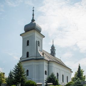 Zdjęcie nr 1: Murowany kościół otrzymał układ jednonawowej świątyni z węższym prezbiterium zwróconym ku północy i wieżą dostawioną do nawy od południa. Prezbiterium jest jednoprzęsłowe zakończone spłaszczoną apsydą. Na jego przedłużeniu usytuowano czworoboczną zakrystię. Prostokątna nawa jest trójprzęsłowa o zaokrąglonych narożnikach. Od frontu wzniesiono czworoboczną wieżę o ściętych narożnikach, flankowaną piętrowymi lokalnościami. Wysunięto ją lekko ku przodowi, gdzie przechodzi wklęsłymi płaszczyznami w boczne części fasady. Jej górne piętro nakryte cebulastym hełmem zostało opięte po bokach wolutami, obok których ustawiono kamienne klasycystyczne wazy. Elewacje zewnętrzne artykułowano podziałami ramowymi, zamykając wysokości ścian gzymsem gurtowy. Na końcu nawy na siodłowym dachu pokrytym blachą umieszczono czworoboczną wieżyczkę na sygnaturkę zwieńczoną cebulastym hełmem z krzyżem. Podobnym dachem nakryto prezbiterium. Wnętrze kościoła zostało rozczłonkowane lizenami z wklęsłymi półlizenami po bokach, ponad którymi rozciągnięto odcinki belkowania. Całość nakryto sklepieniem żaglastym na zdwojonych gurtach. Od południa w nawie urządzono chór muzyczny wsparty na dwóch kolumnach z parapetem wklęsłowypukłym. W prezbiterium usytuowano ołtarz główny, a w nawie dwa ołtarze boczne. Tu także na ścianie zachodniej zawieszono ambonę.  