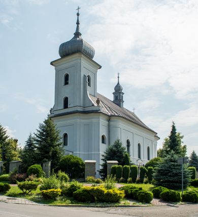 Zdjęcie nr 2: Murowany kościół otrzymał układ jednonawowej świątyni z węższym prezbiterium zwróconym ku północy i wieżą dostawioną do nawy od południa. Prezbiterium jest jednoprzęsłowe zakończone spłaszczoną apsydą. Na jego przedłużeniu usytuowano czworoboczną zakrystię. Prostokątna nawa jest trójprzęsłowa o zaokrąglonych narożnikach. Od frontu wzniesiono czworoboczną wieżę o ściętych narożnikach, flankowaną piętrowymi lokalnościami. Wysunięto ją lekko ku przodowi, gdzie przechodzi wklęsłymi płaszczyznami w boczne części fasady. Jej górne piętro nakryte cebulastym hełmem zostało opięte po bokach wolutami, obok których ustawiono kamienne klasycystyczne wazy. Elewacje zewnętrzne artykułowano podziałami ramowymi, zamykając wysokości ścian gzymsem gurtowy. Na końcu nawy na siodłowym dachu pokrytym blachą umieszczono czworoboczną wieżyczkę na sygnaturkę zwieńczoną cebulastym hełmem z krzyżem. Podobnym dachem nakryto prezbiterium. Wnętrze kościoła zostało rozczłonkowane lizenami z wklęsłymi półlizenami po bokach, ponad którymi rozciągnięto odcinki belkowania. Całość nakryto sklepieniem żaglastym na zdwojonych gurtach. Od południa w nawie urządzono chór muzyczny wsparty na dwóch kolumnach z parapetem wklęsłowypukłym. W prezbiterium usytuowano ołtarz główny, a w nawie dwa ołtarze boczne. Tu także na ścianie zachodniej zawieszono ambonę.  