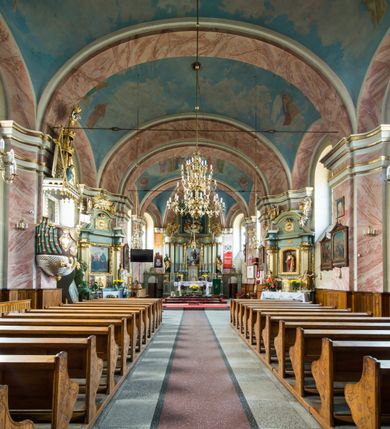 Zdjęcie nr 6: Murowany kościół otrzymał układ jednonawowej świątyni z węższym prezbiterium zwróconym ku północy i wieżą dostawioną do nawy od południa. Prezbiterium jest jednoprzęsłowe zakończone spłaszczoną apsydą. Na jego przedłużeniu usytuowano czworoboczną zakrystię. Prostokątna nawa jest trójprzęsłowa o zaokrąglonych narożnikach. Od frontu wzniesiono czworoboczną wieżę o ściętych narożnikach, flankowaną piętrowymi lokalnościami. Wysunięto ją lekko ku przodowi, gdzie przechodzi wklęsłymi płaszczyznami w boczne części fasady. Jej górne piętro nakryte cebulastym hełmem zostało opięte po bokach wolutami, obok których ustawiono kamienne klasycystyczne wazy. Elewacje zewnętrzne artykułowano podziałami ramowymi, zamykając wysokości ścian gzymsem gurtowy. Na końcu nawy na siodłowym dachu pokrytym blachą umieszczono czworoboczną wieżyczkę na sygnaturkę zwieńczoną cebulastym hełmem z krzyżem. Podobnym dachem nakryto prezbiterium. Wnętrze kościoła zostało rozczłonkowane lizenami z wklęsłymi półlizenami po bokach, ponad którymi rozciągnięto odcinki belkowania. Całość nakryto sklepieniem żaglastym na zdwojonych gurtach. Od południa w nawie urządzono chór muzyczny wsparty na dwóch kolumnach z parapetem wklęsłowypukłym. W prezbiterium usytuowano ołtarz główny, a w nawie dwa ołtarze boczne. Tu także na ścianie zachodniej zawieszono ambonę.  