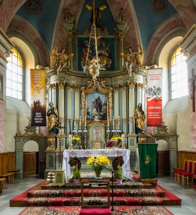 Zdjęcie nr 10: Murowany kościół otrzymał układ jednonawowej świątyni z węższym prezbiterium zwróconym ku północy i wieżą dostawioną do nawy od południa. Prezbiterium jest jednoprzęsłowe zakończone spłaszczoną apsydą. Na jego przedłużeniu usytuowano czworoboczną zakrystię. Prostokątna nawa jest trójprzęsłowa o zaokrąglonych narożnikach. Od frontu wzniesiono czworoboczną wieżę o ściętych narożnikach, flankowaną piętrowymi lokalnościami. Wysunięto ją lekko ku przodowi, gdzie przechodzi wklęsłymi płaszczyznami w boczne części fasady. Jej górne piętro nakryte cebulastym hełmem zostało opięte po bokach wolutami, obok których ustawiono kamienne klasycystyczne wazy. Elewacje zewnętrzne artykułowano podziałami ramowymi, zamykając wysokości ścian gzymsem gurtowy. Na końcu nawy na siodłowym dachu pokrytym blachą umieszczono czworoboczną wieżyczkę na sygnaturkę zwieńczoną cebulastym hełmem z krzyżem. Podobnym dachem nakryto prezbiterium. Wnętrze kościoła zostało rozczłonkowane lizenami z wklęsłymi półlizenami po bokach, ponad którymi rozciągnięto odcinki belkowania. Całość nakryto sklepieniem żaglastym na zdwojonych gurtach. Od południa w nawie urządzono chór muzyczny wsparty na dwóch kolumnach z parapetem wklęsłowypukłym. W prezbiterium usytuowano ołtarz główny, a w nawie dwa ołtarze boczne. Tu także na ścianie zachodniej zawieszono ambonę.  